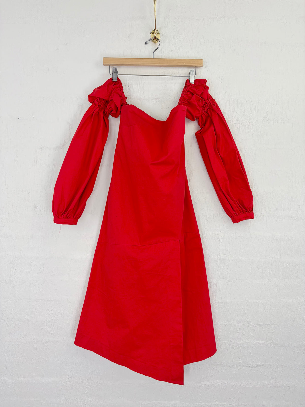 A bright red off-the-shoulder dress with long balloon sleeves, hanging from a wooden hanger against a white painted brick wall.