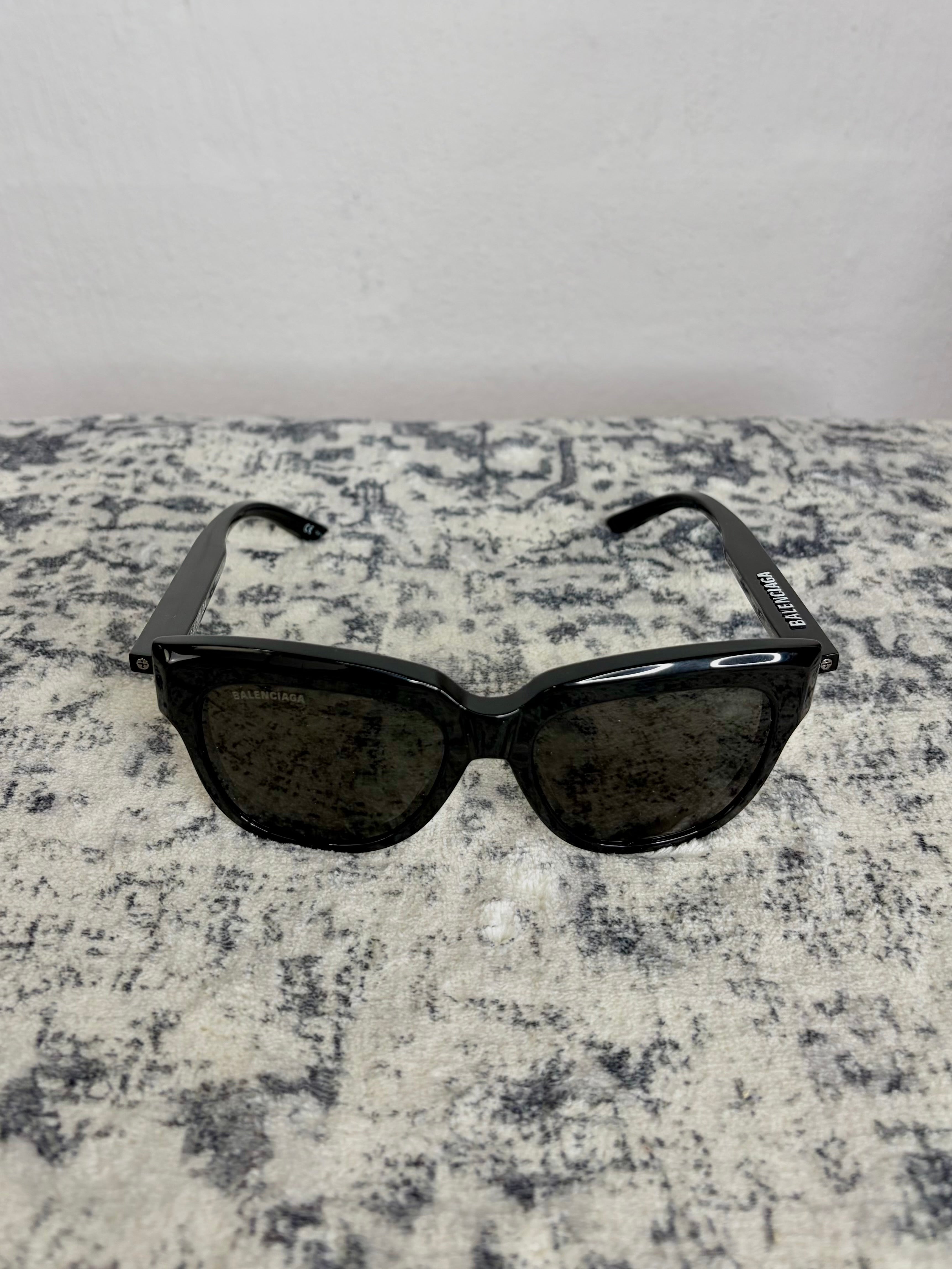 Black cat-eye sunglasses with dark tinted lenses, displayed on a textured gray and white surface against a light gray wall.