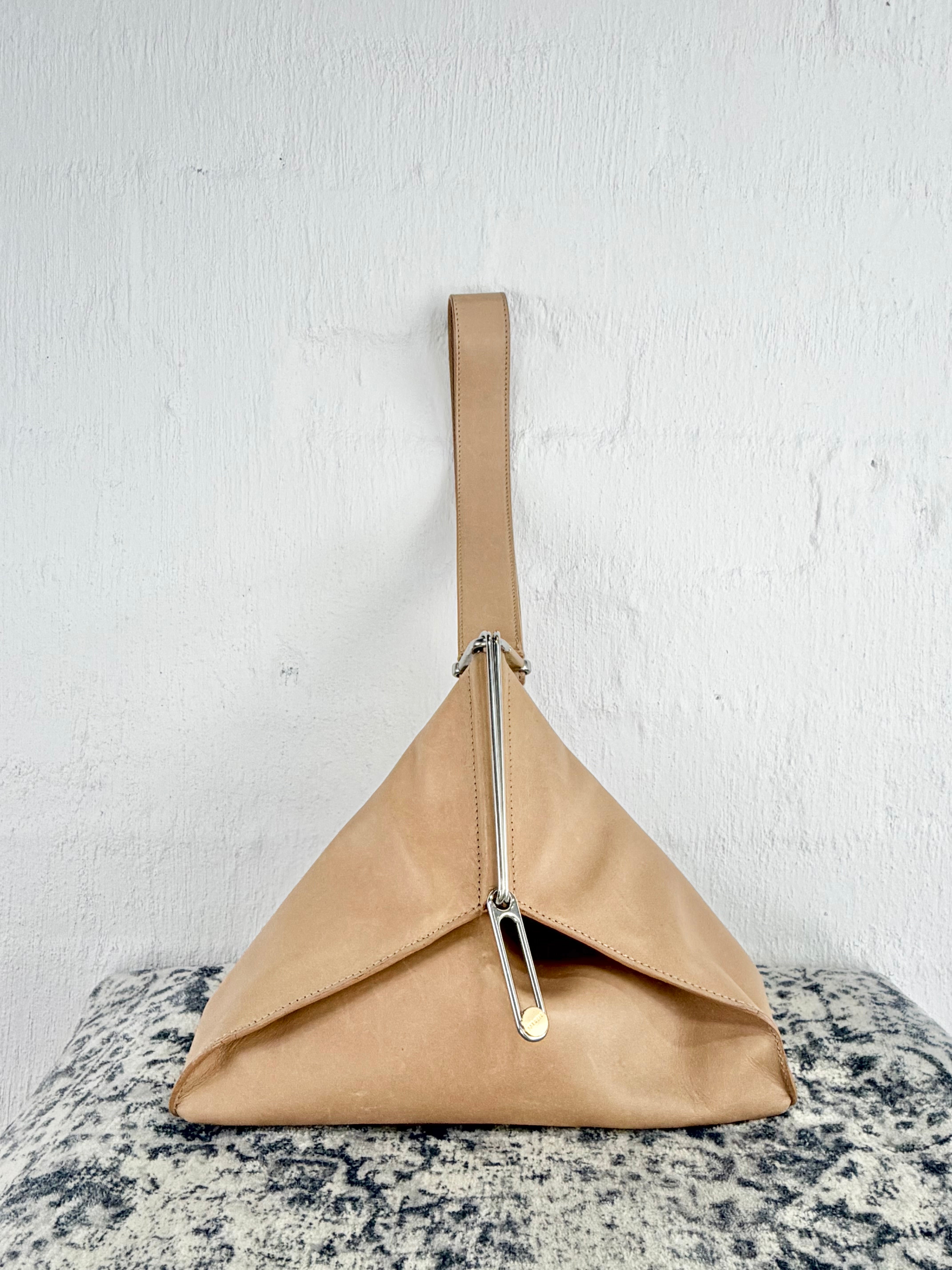 A structured, pyramid-shaped shoulder bag in tan leather with a silver-tone safety pin detail and strap, resting on a textured gray and white surface against a white wall.