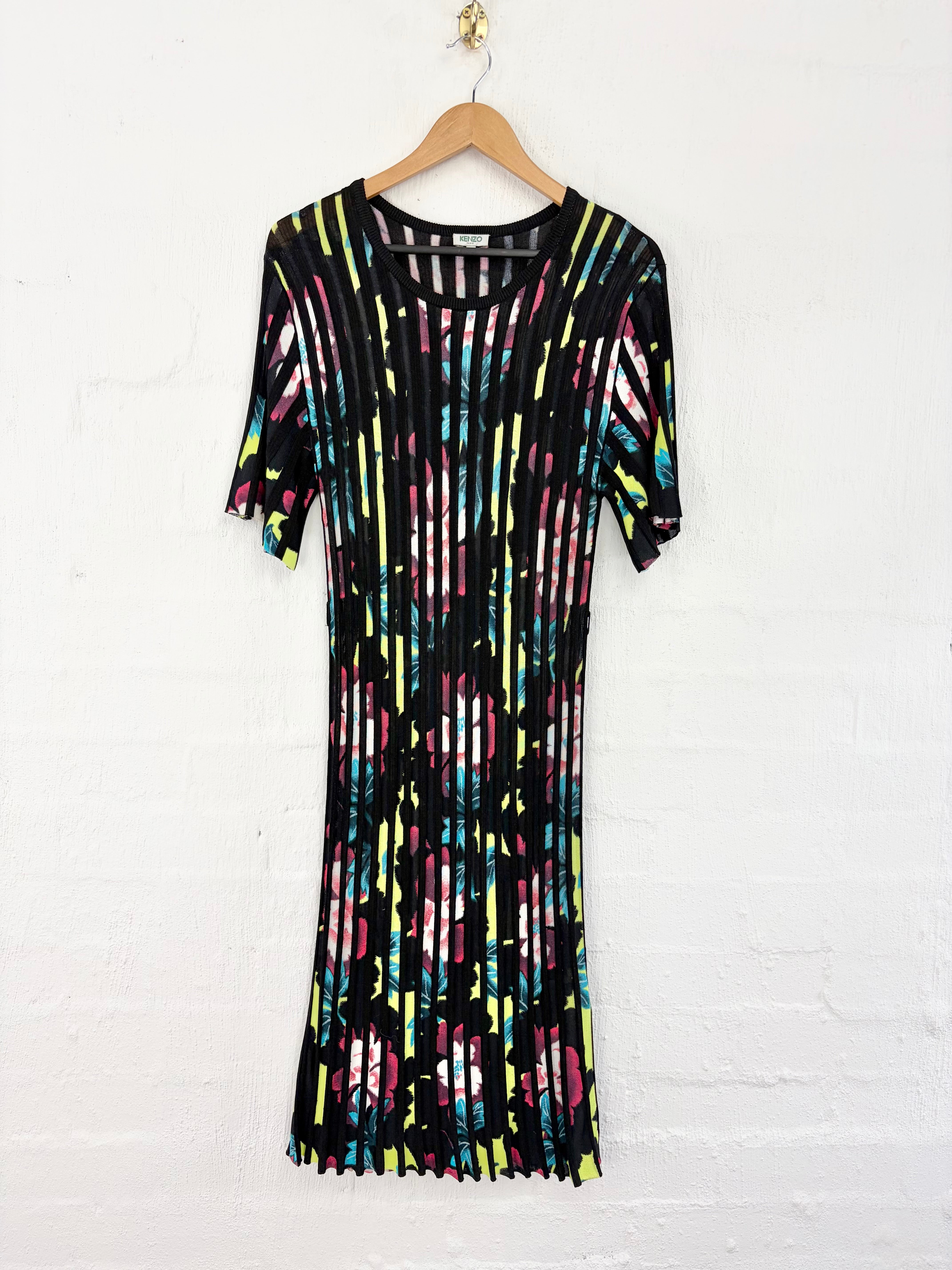 A short-sleeve midi dress with a black base and a vibrant multicolor vertical stripe pattern, hanging on a wooden hanger against a white painted brick wall.
