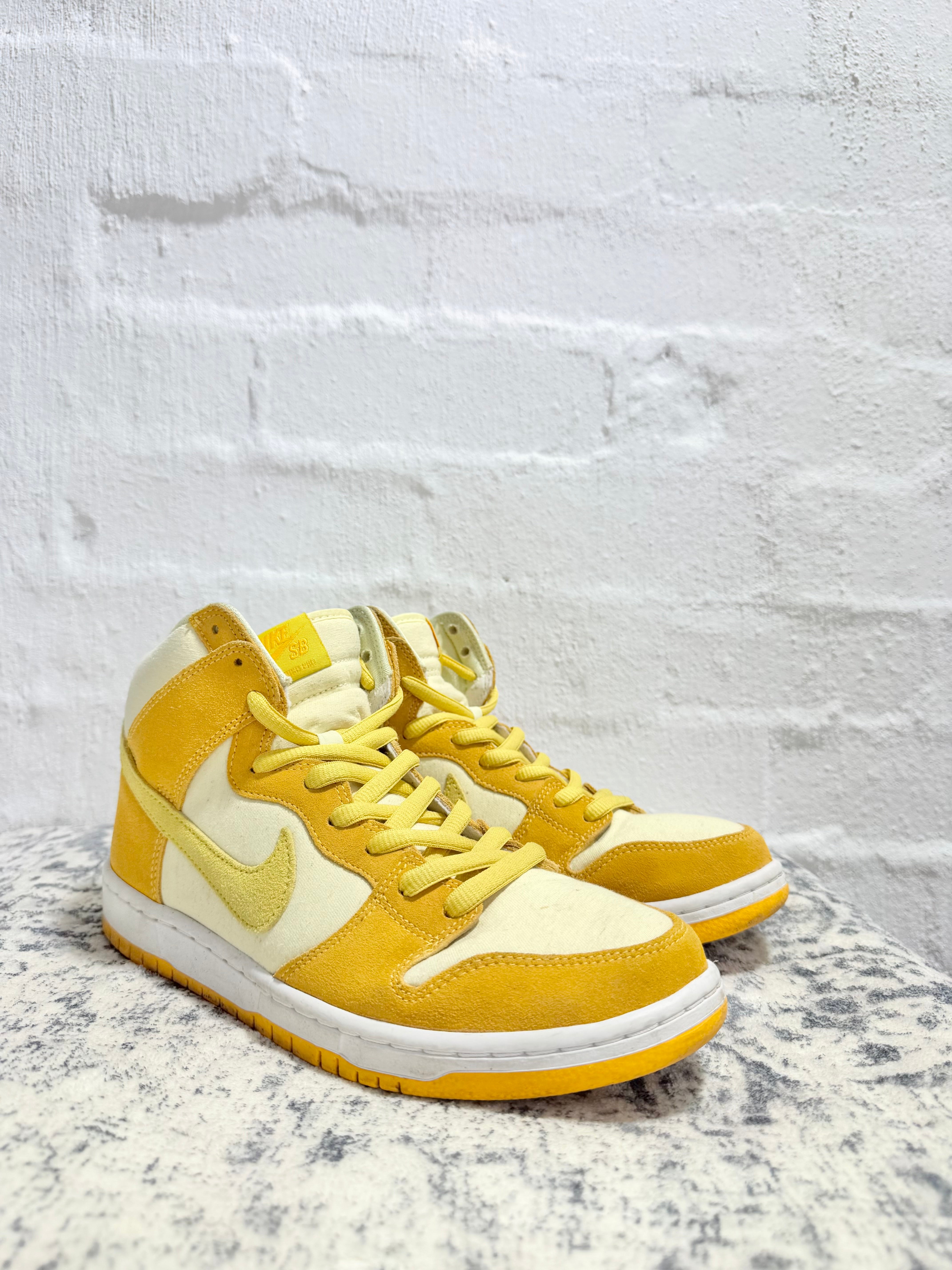 A pair of Nike high-top sneakers in a yellow and cream colorway, displayed on a textured gray surface against a white brick wall background.