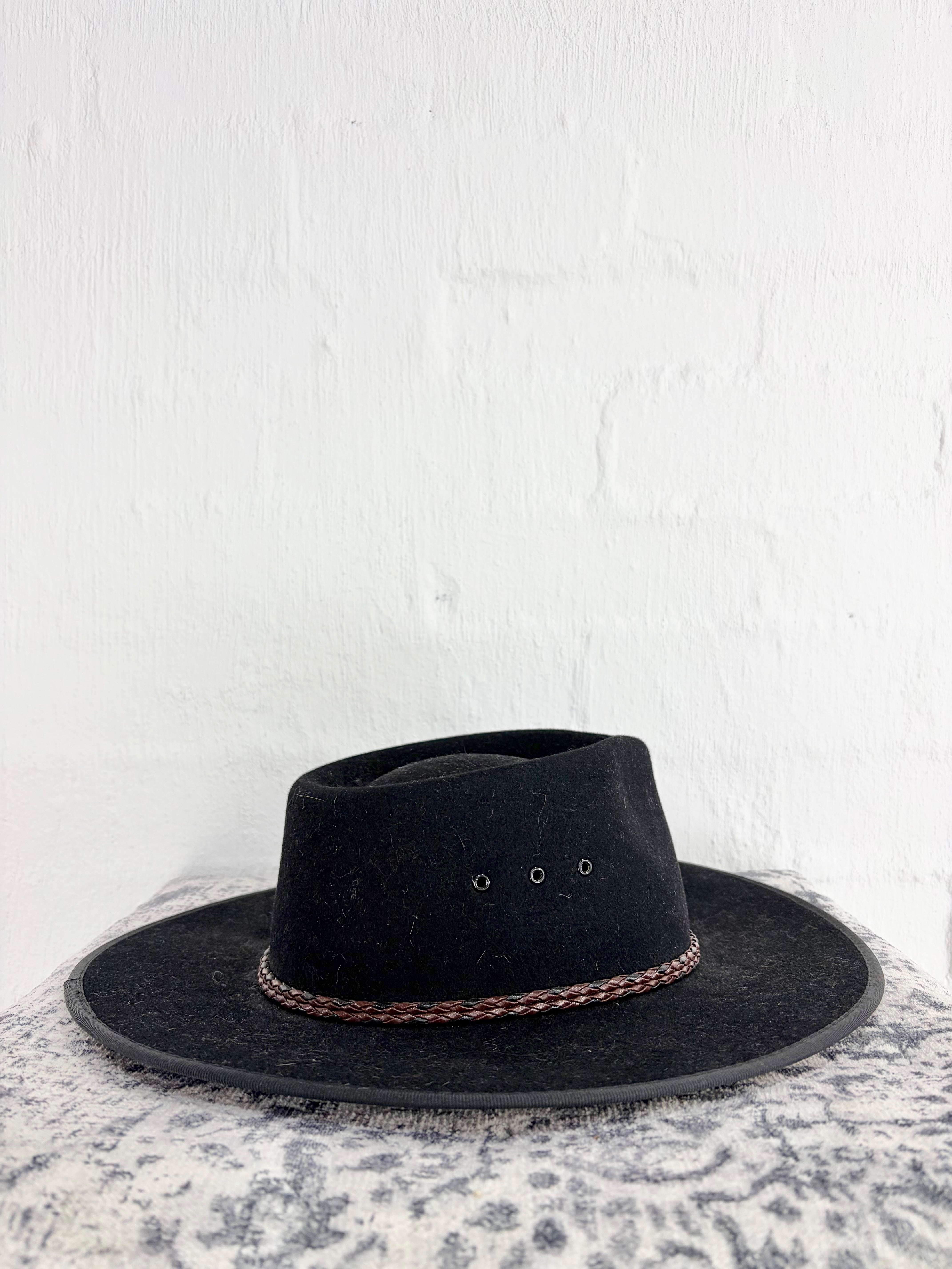 A black wide-brim fedora hat with a brown braided band, displayed on a textured gray and white surface against a white brick wall.