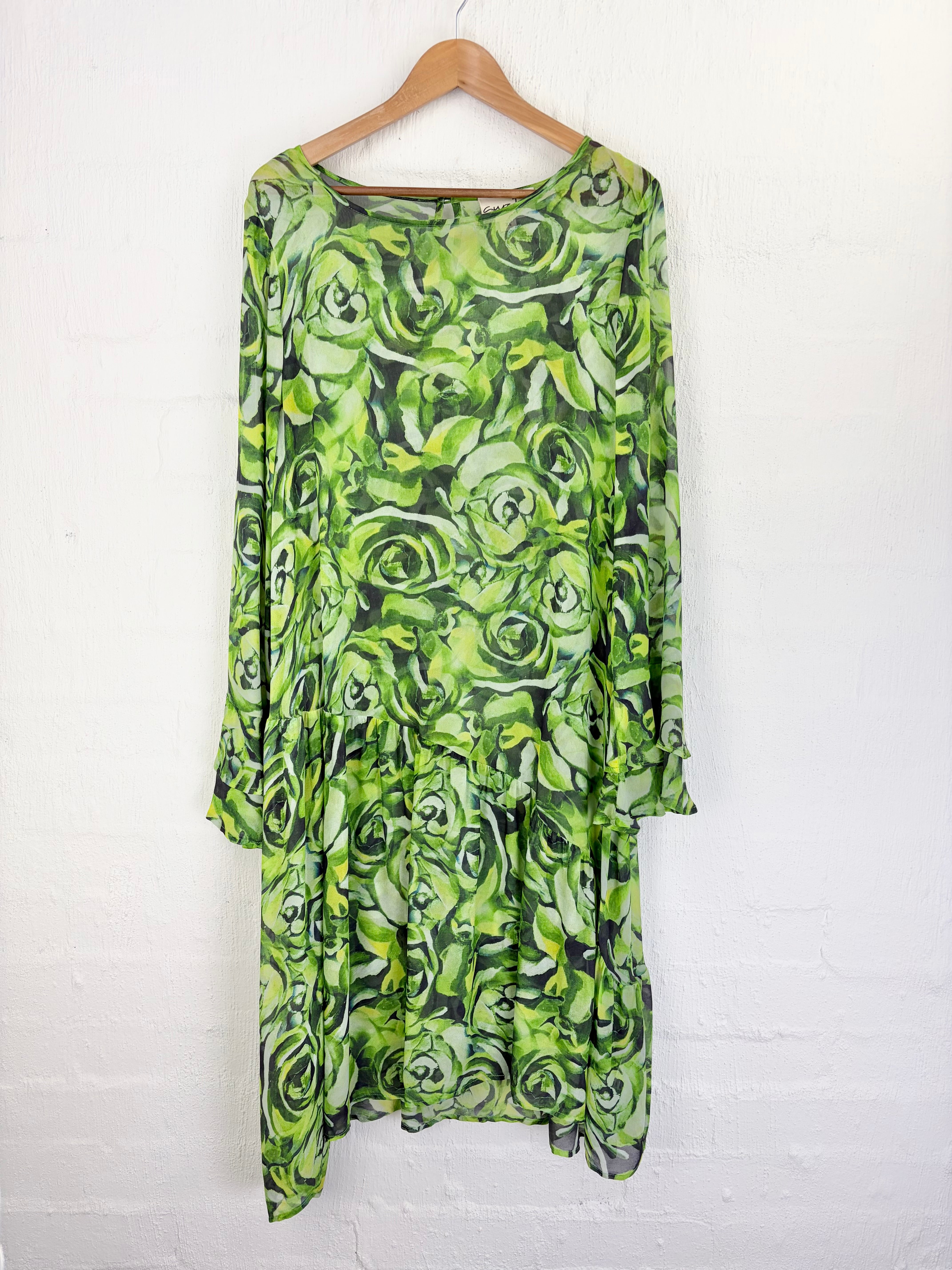 A long-sleeve midi dress with an all-over green rose print, hanging on a wooden hanger against a white painted brick wall.