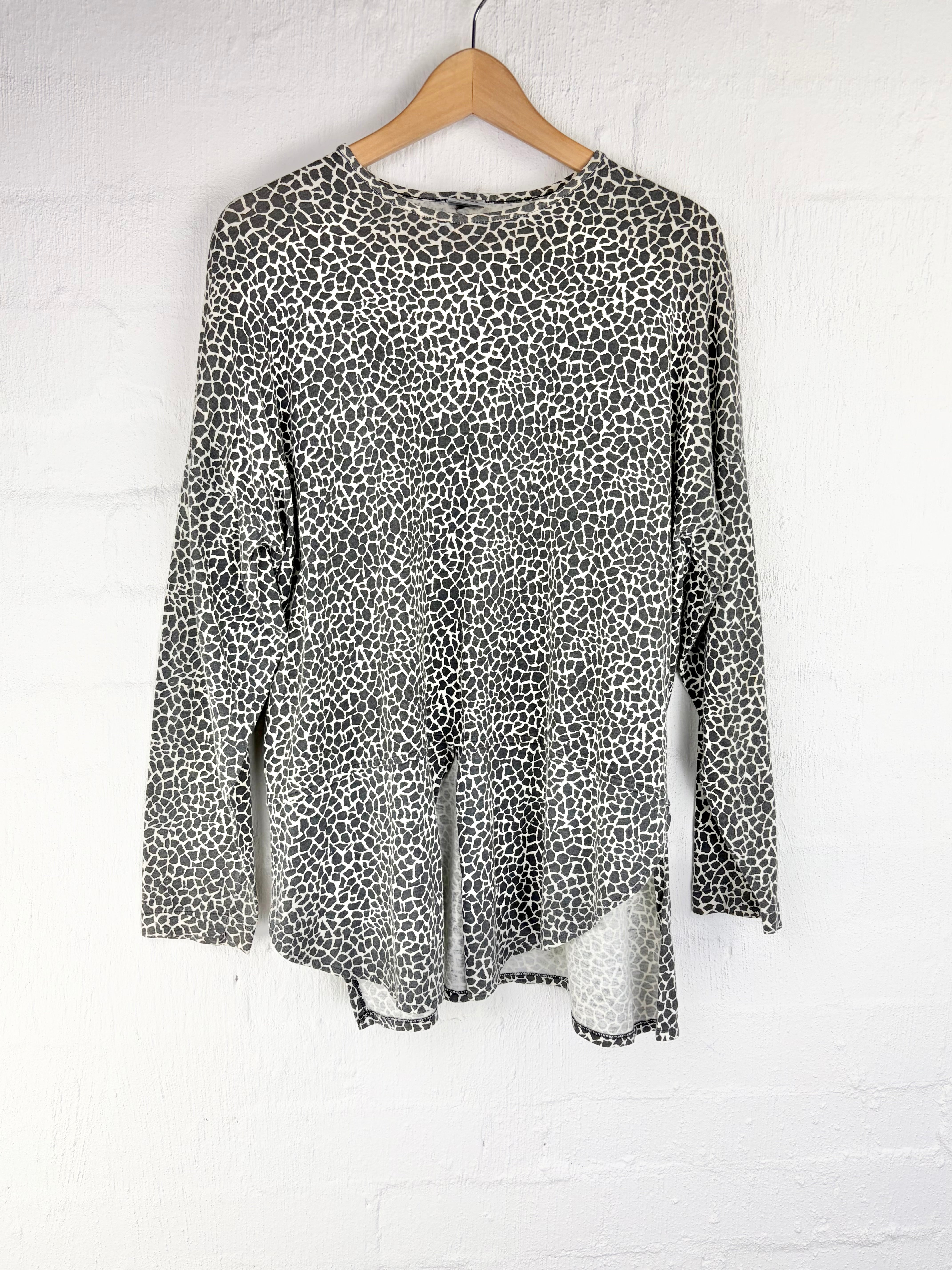 A long-sleeve tunic top with a black and white leopard print pattern, hanging on a wooden hanger against a white painted brick wall.