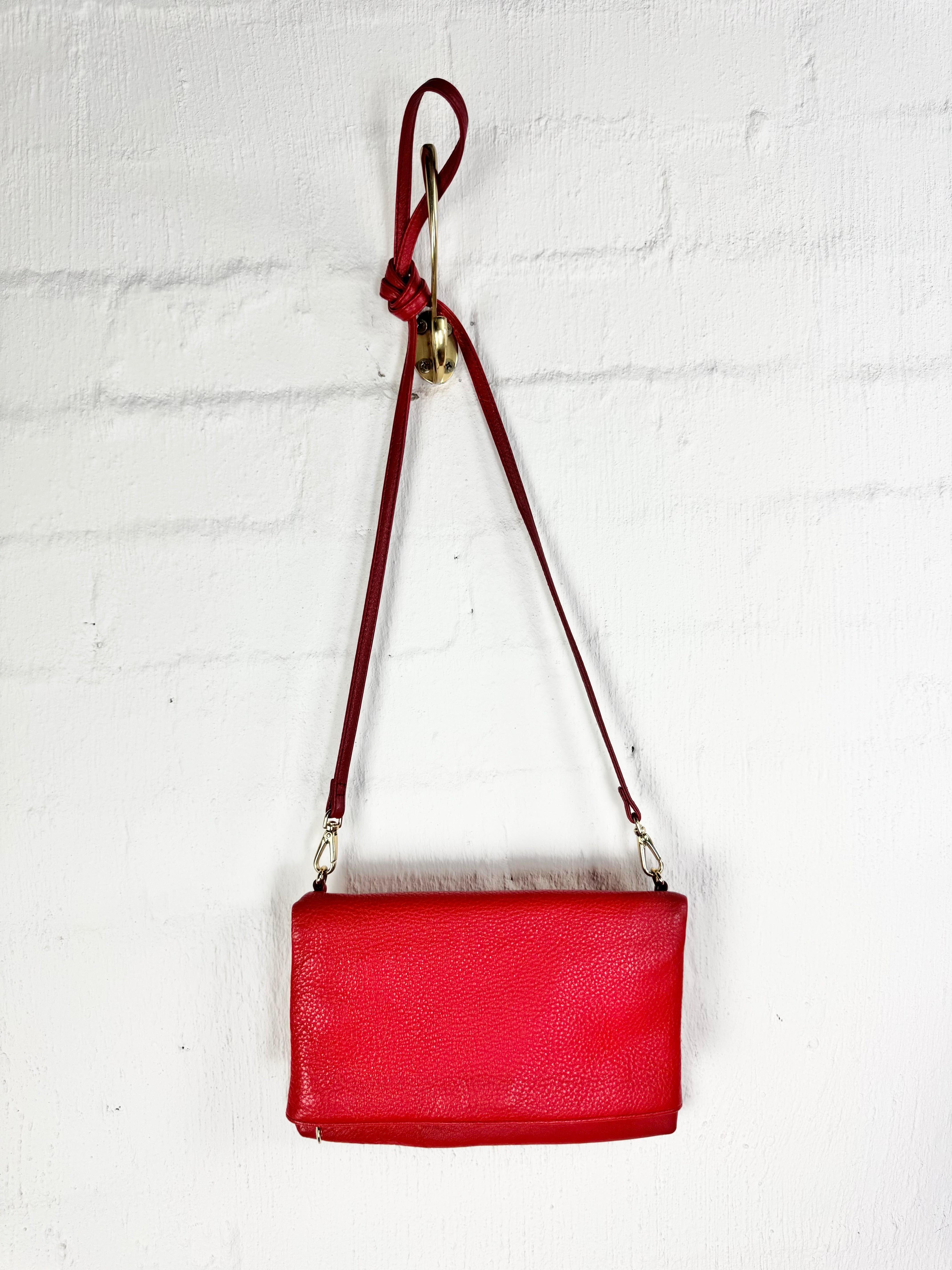 A red pebbled leather crossbody bag with a matching red strap and gold-tone hardware, hanging against a white painted brick wall.
