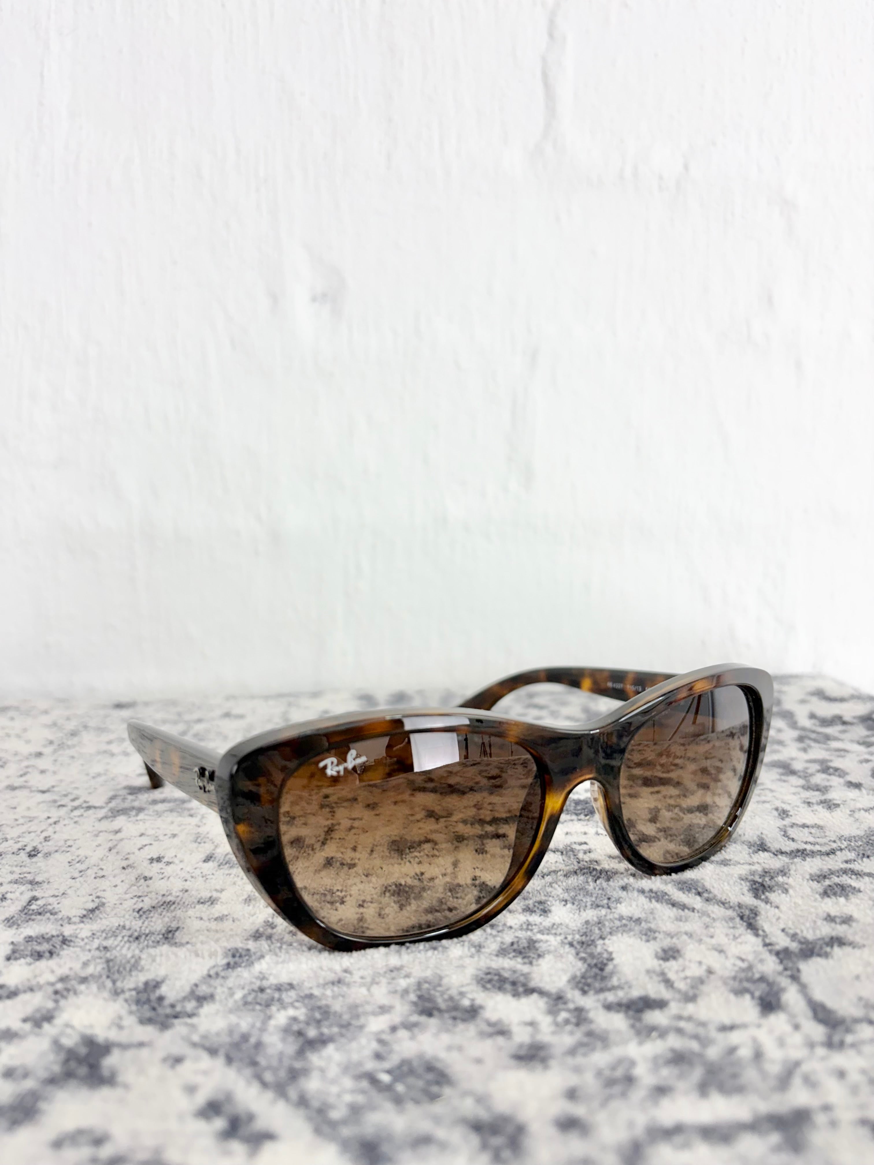 A pair of Ray-Ban sunglasses with a brown tortoiseshell acetate frame and gradient brown lenses, resting on a textured gray and white surface against a white brick wall.