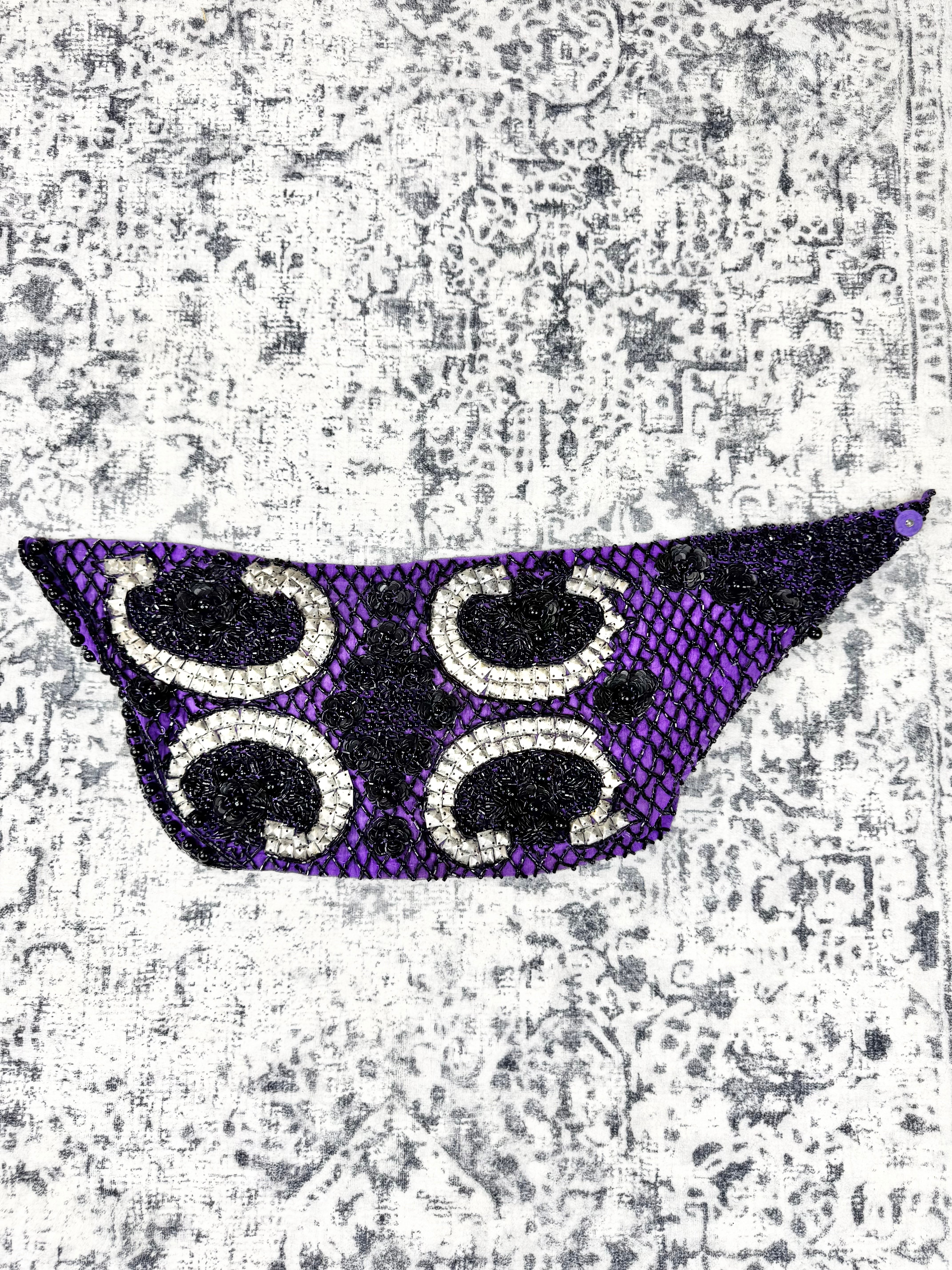 A purple and black beaded mask-shaped accessory with four white and black paisley-like bead motifs, displayed on a distressed gray and white patterned background.
