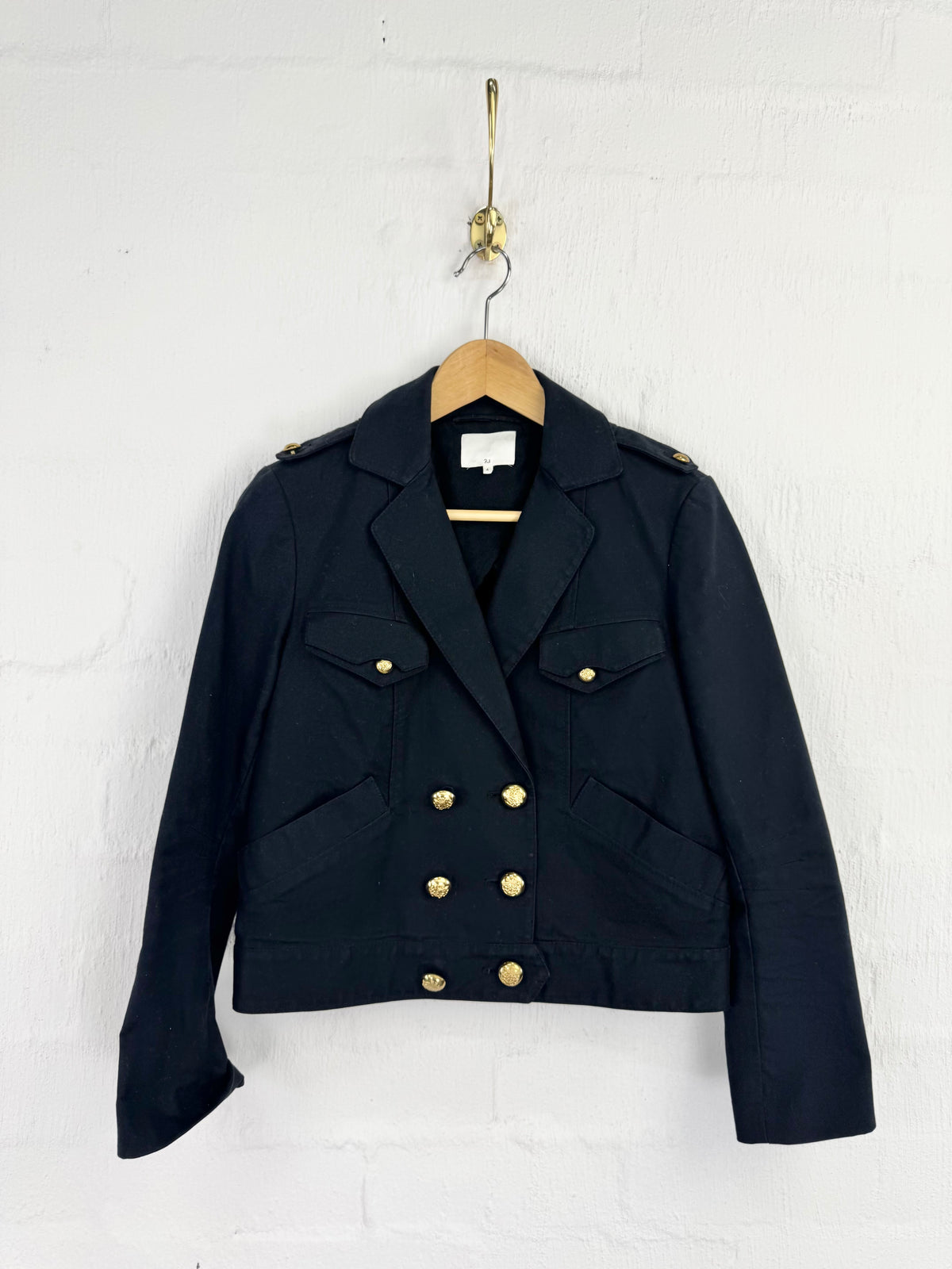 A black double-breasted blazer jacket with gold buttons and shoulder epaulets, hanging on a wooden hanger against a white painted brick wall.