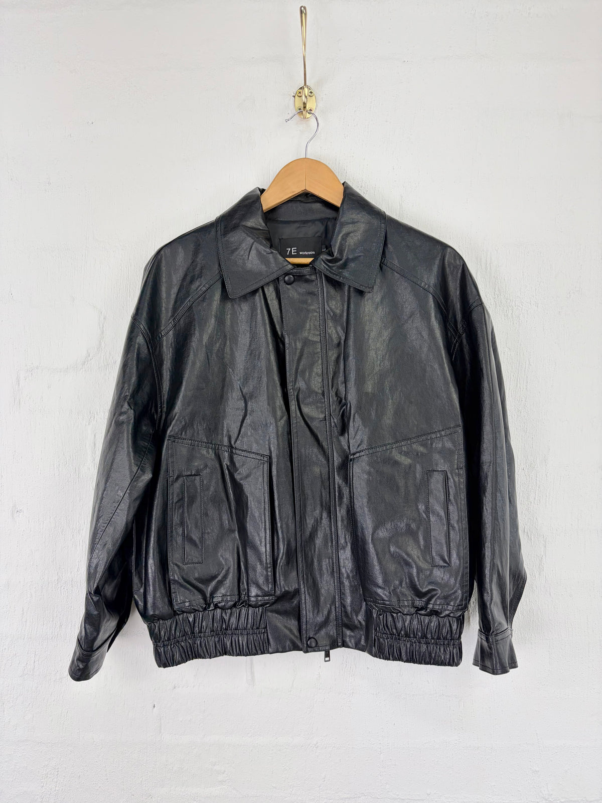 A black leather jacket hanging on a wooden hanger against a white painted brick wall.