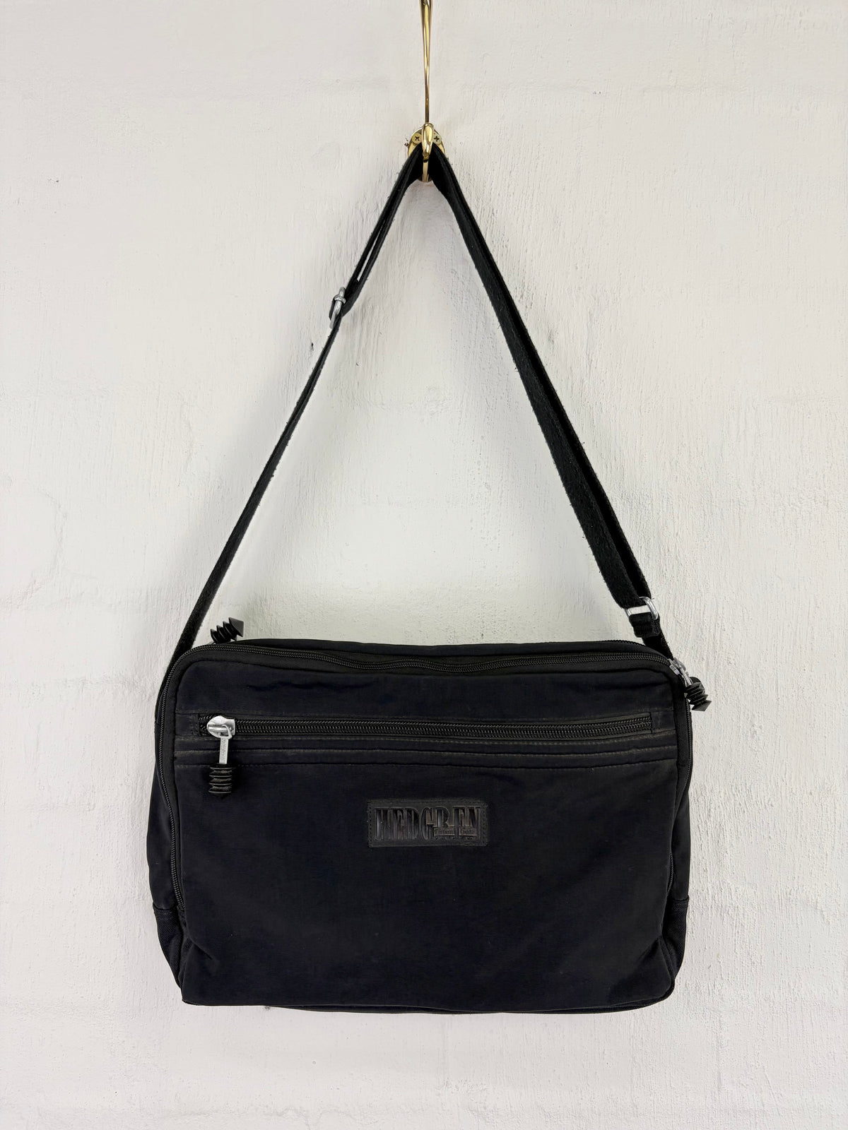 A black nylon shoulder bag with a front zippered pocket and a small 'TWENTYSEVEN' brand patch, hanging from a gold hook against a white brick wall.