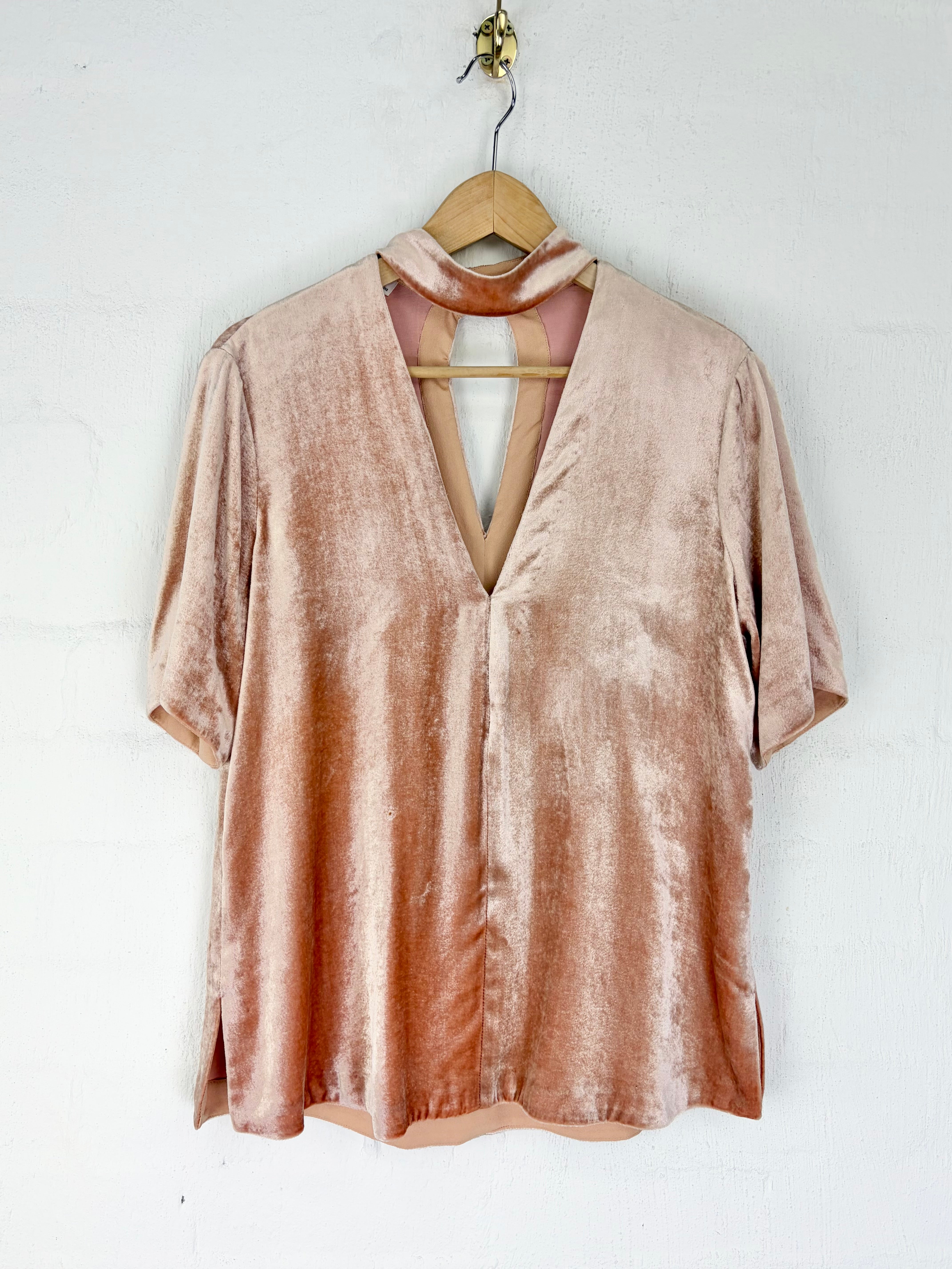 A short-sleeve dusty rose velvet top with a keyhole cutout and tie detail, hanging on a wooden hanger against a white wall.