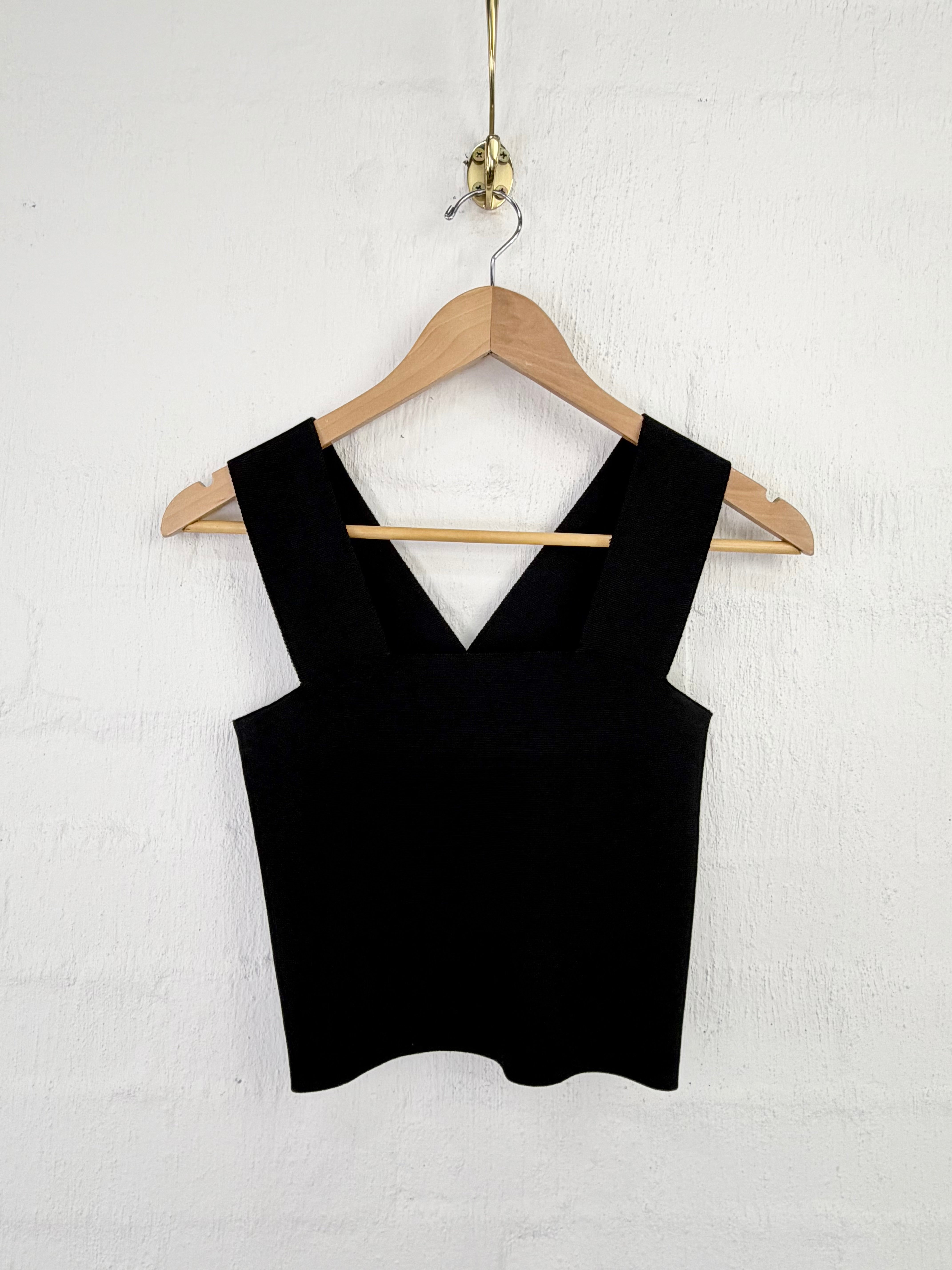 A black sleeveless knit top with wide straps and a V-neckline, hanging on a wooden hanger against a white textured wall.