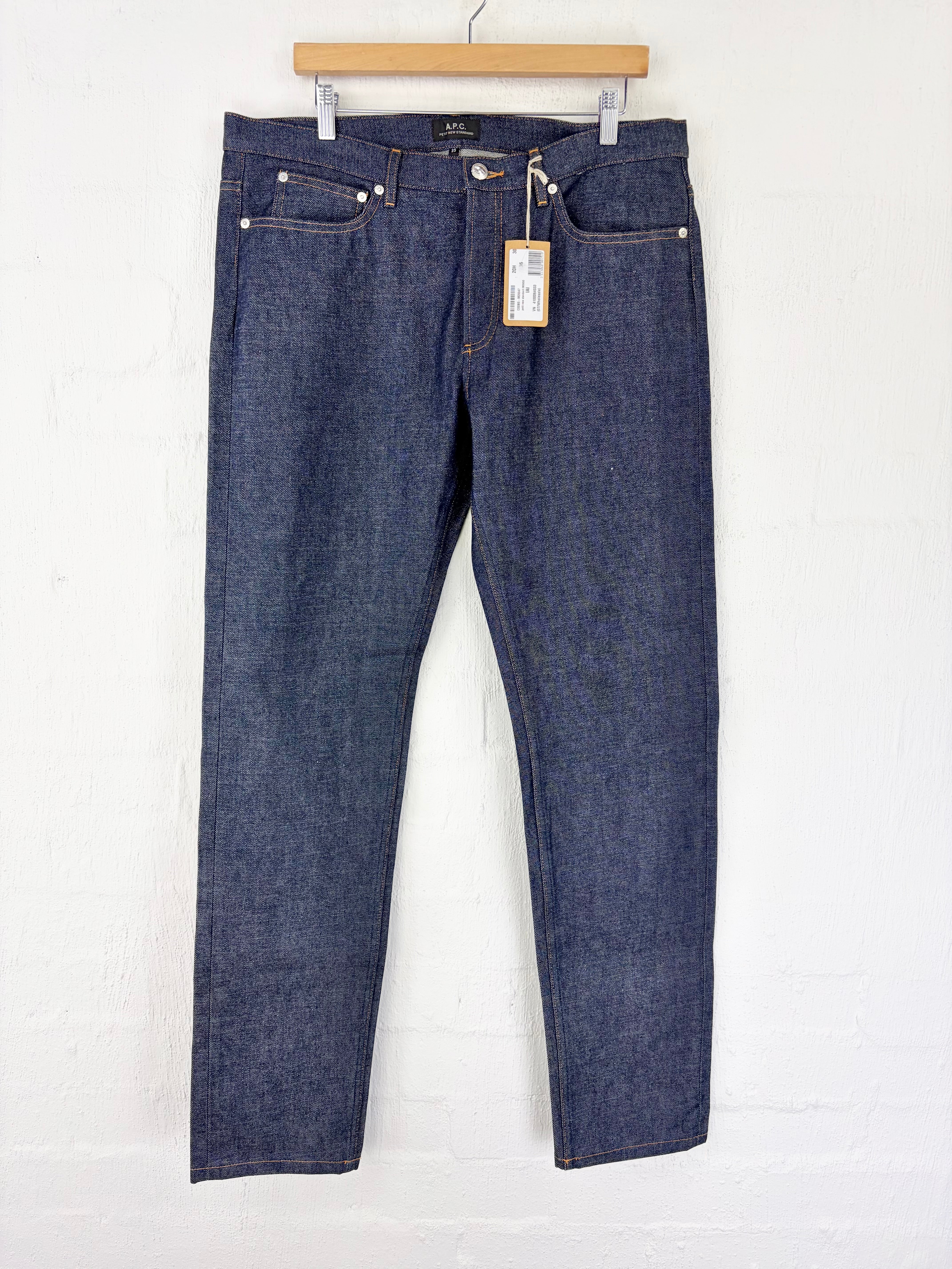 A pair of dark blue denim jeans hanging from a wooden hanger against a white painted brick wall. The jeans have silver rivets, a light wood fly button, and a paper price tag hanging from the waistband.