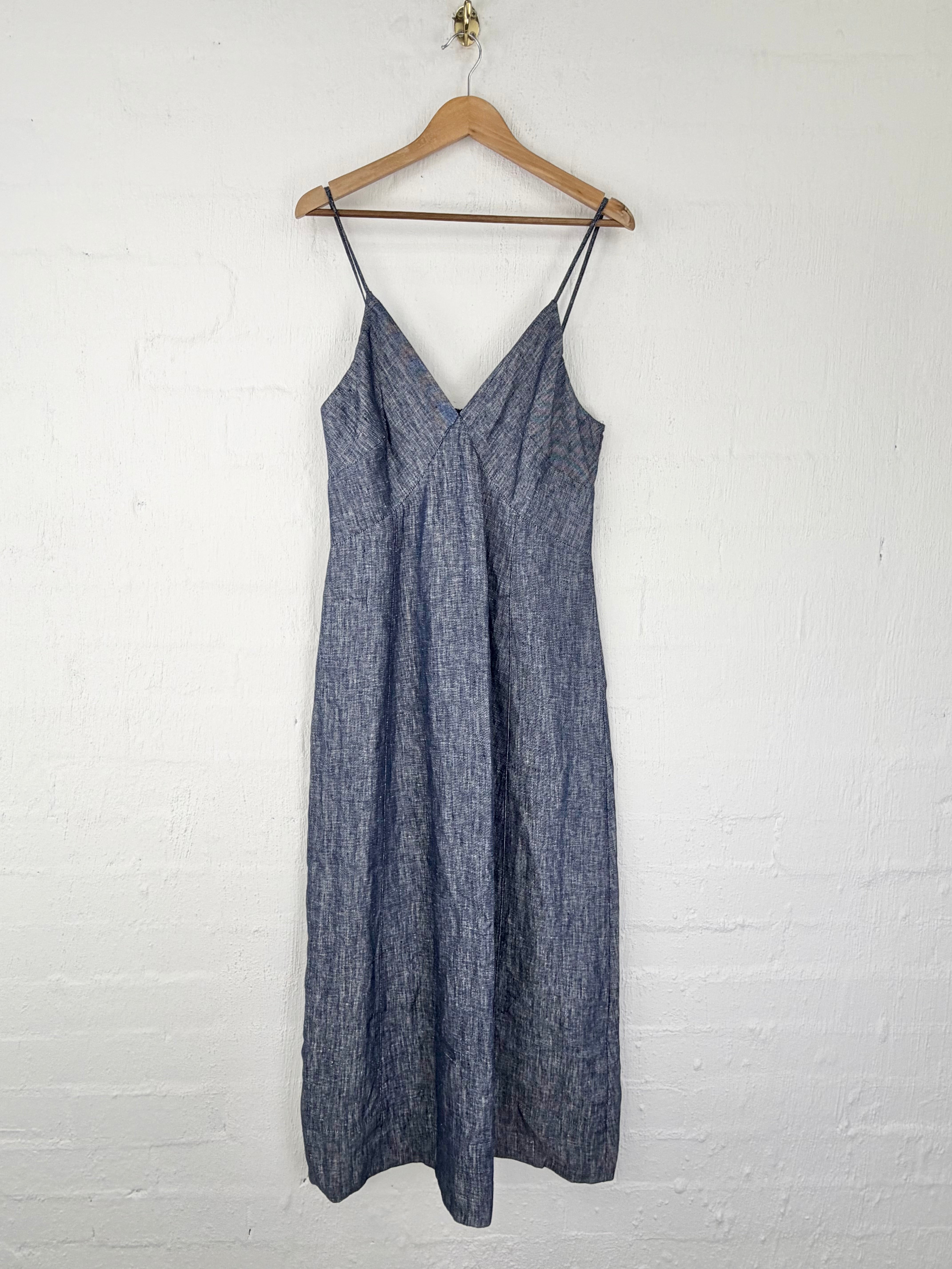A sleeveless blue-gray textured slip dress with thin spaghetti straps and a V-neckline, hanging on a wooden hanger against a white painted brick wall.