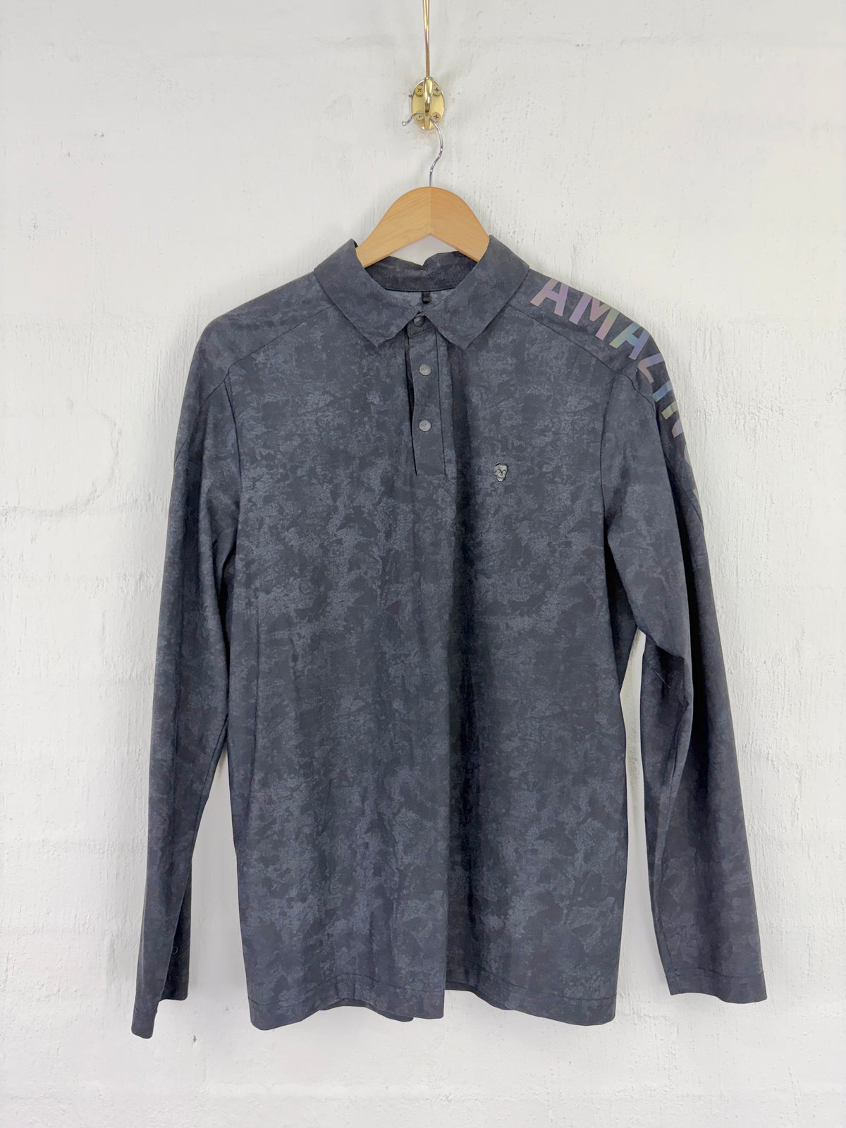 A dark gray long-sleeve polo shirt with a subtle textured pattern, hanging on a wooden hanger against a white brick wall. The right shoulder features a colorful graphic print with the letters 'AWKWARD' in a multicolor pattern.