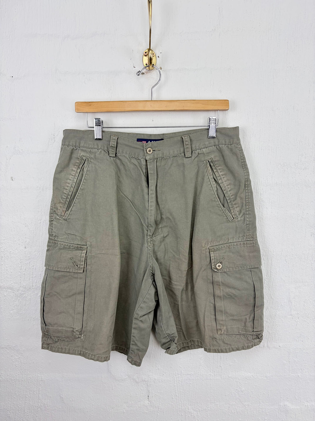 Olive green cargo shorts hanging from a wooden hanger against a white painted brick wall.