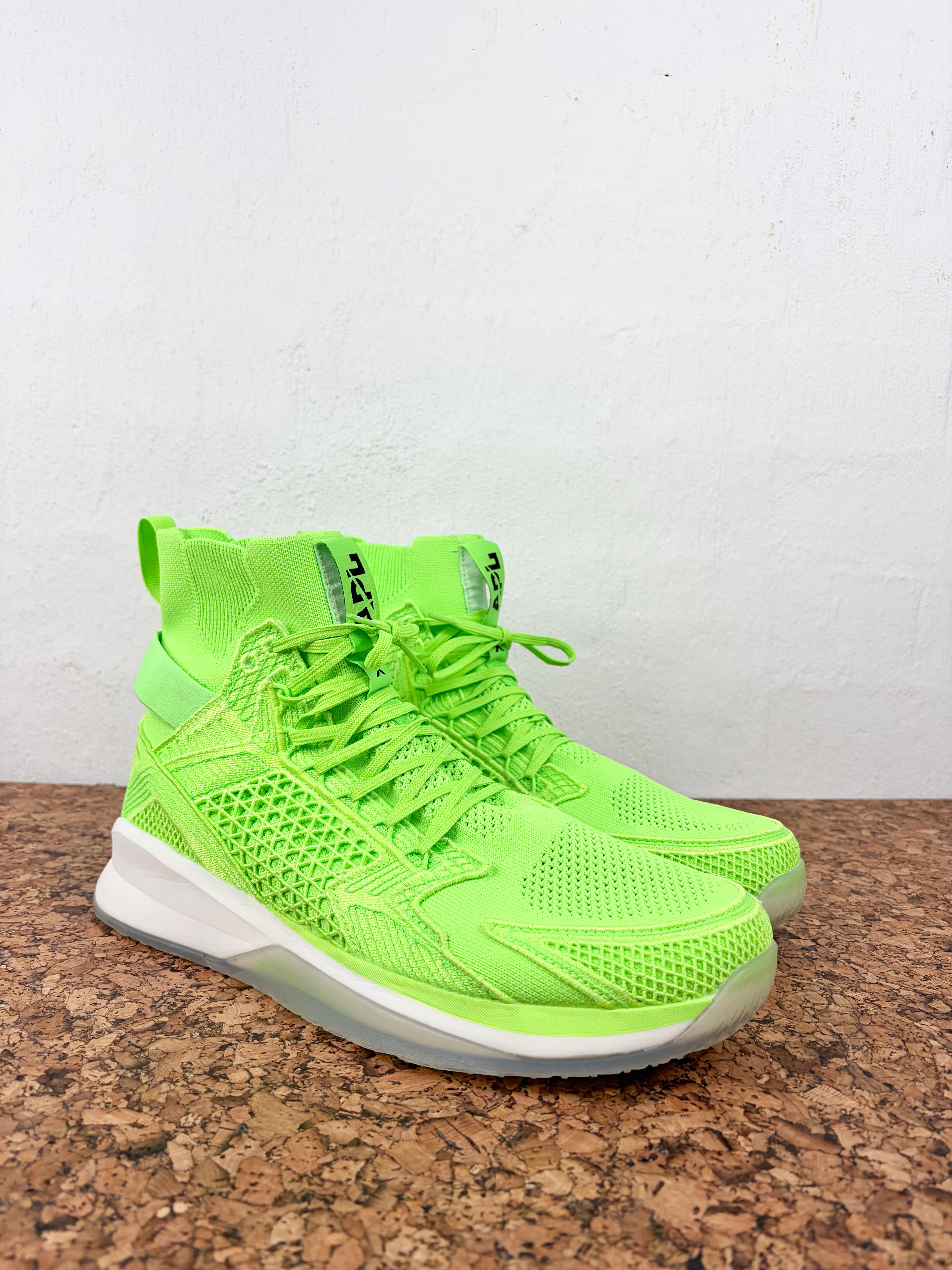 A pair of neon green athletic sneakers with a knit upper, white midsoles, and clear outsoles, displayed on a brown speckled surface against a white wall.