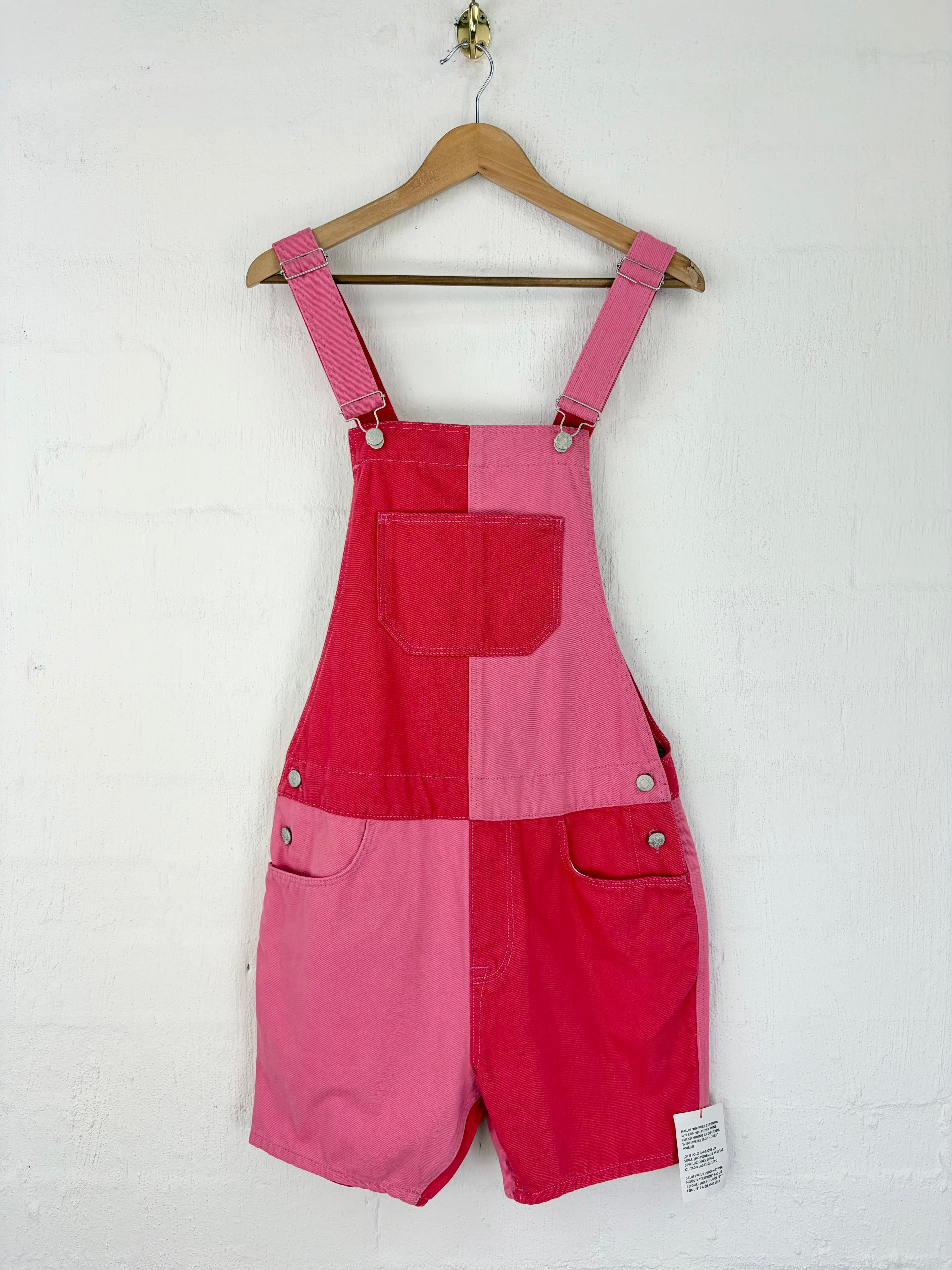 Pink patchwork short overalls hanging on a wooden hanger against a white textured wall.