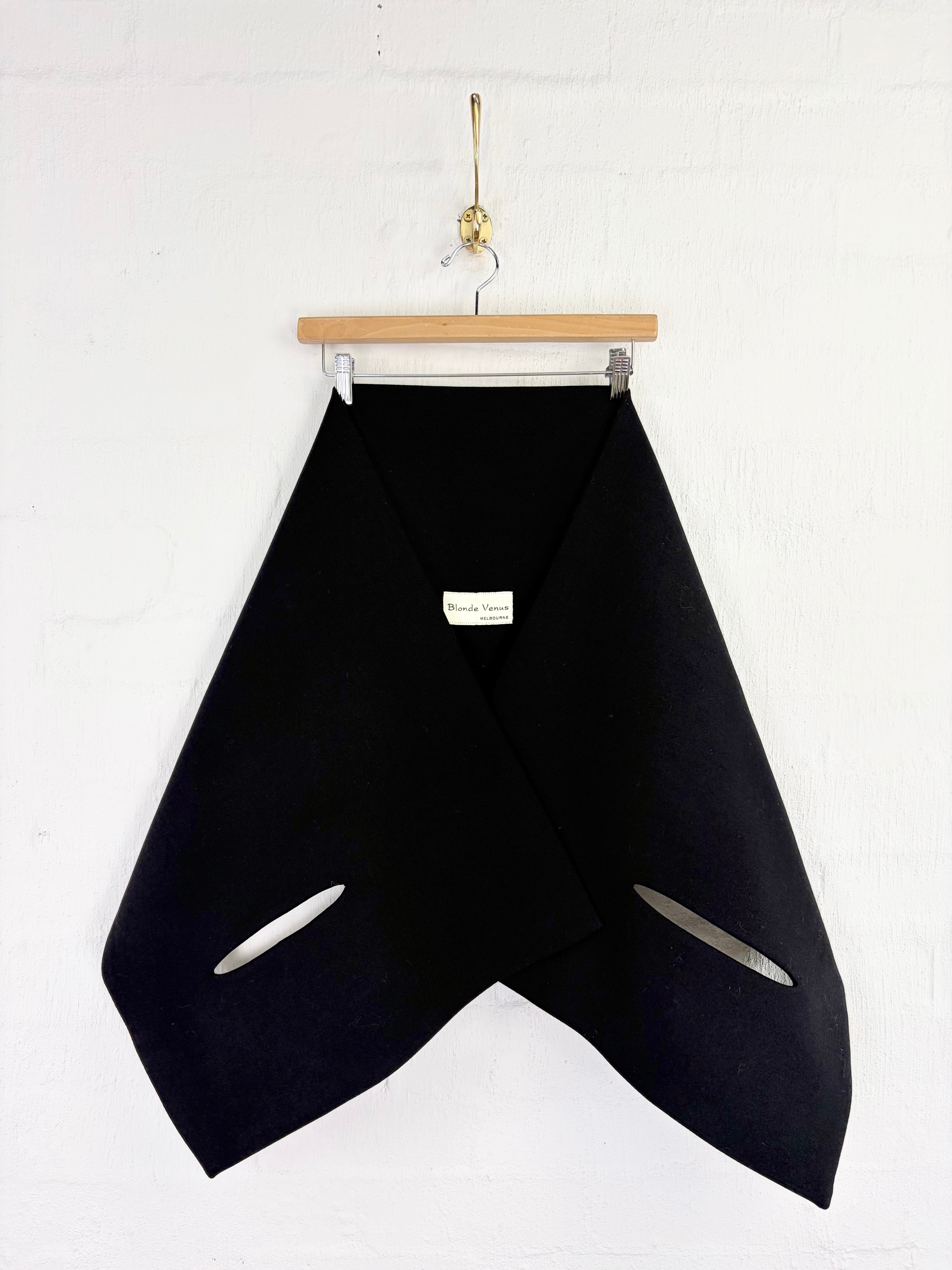 A black asymmetrical wrap jacket with diagonal cutouts, hanging from a wooden hanger against a white painted brick wall.