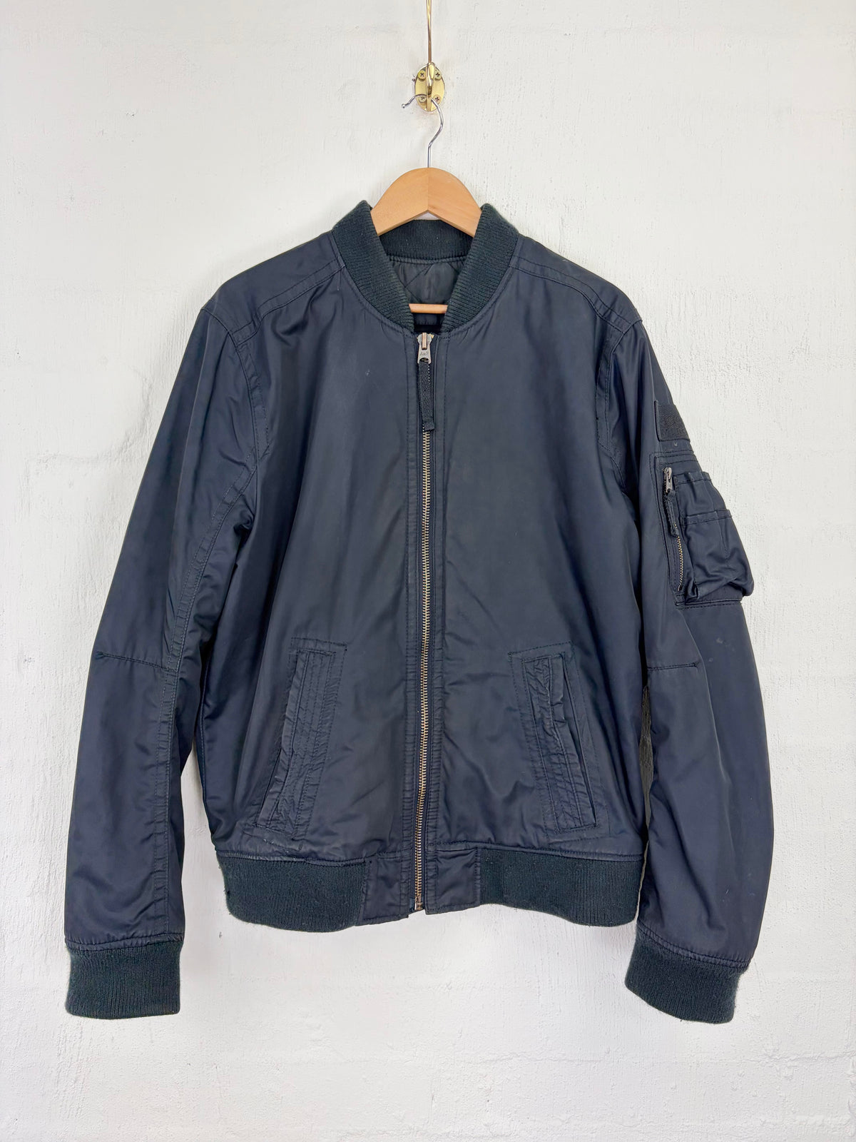 A dark navy blue bomber jacket hanging on a wooden hanger against a white painted brick wall. The jacket features a front zipper, ribbed collar, cuffs, and hem, and a zippered sleeve pocket.
