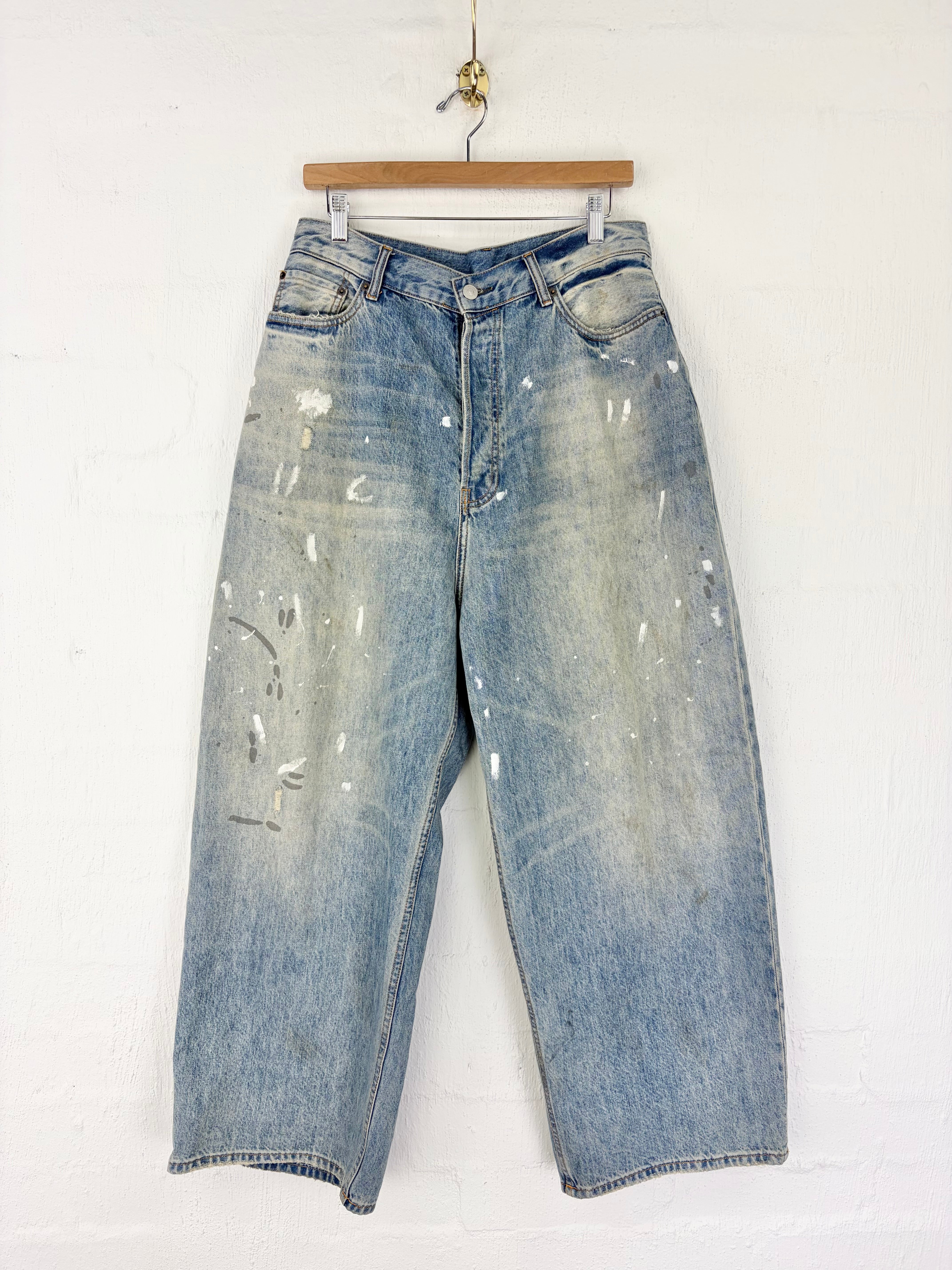 Light blue washed denim cropped pants with distressed details and white paint splatter accents, hanging on a wooden hanger against a white brick wall.