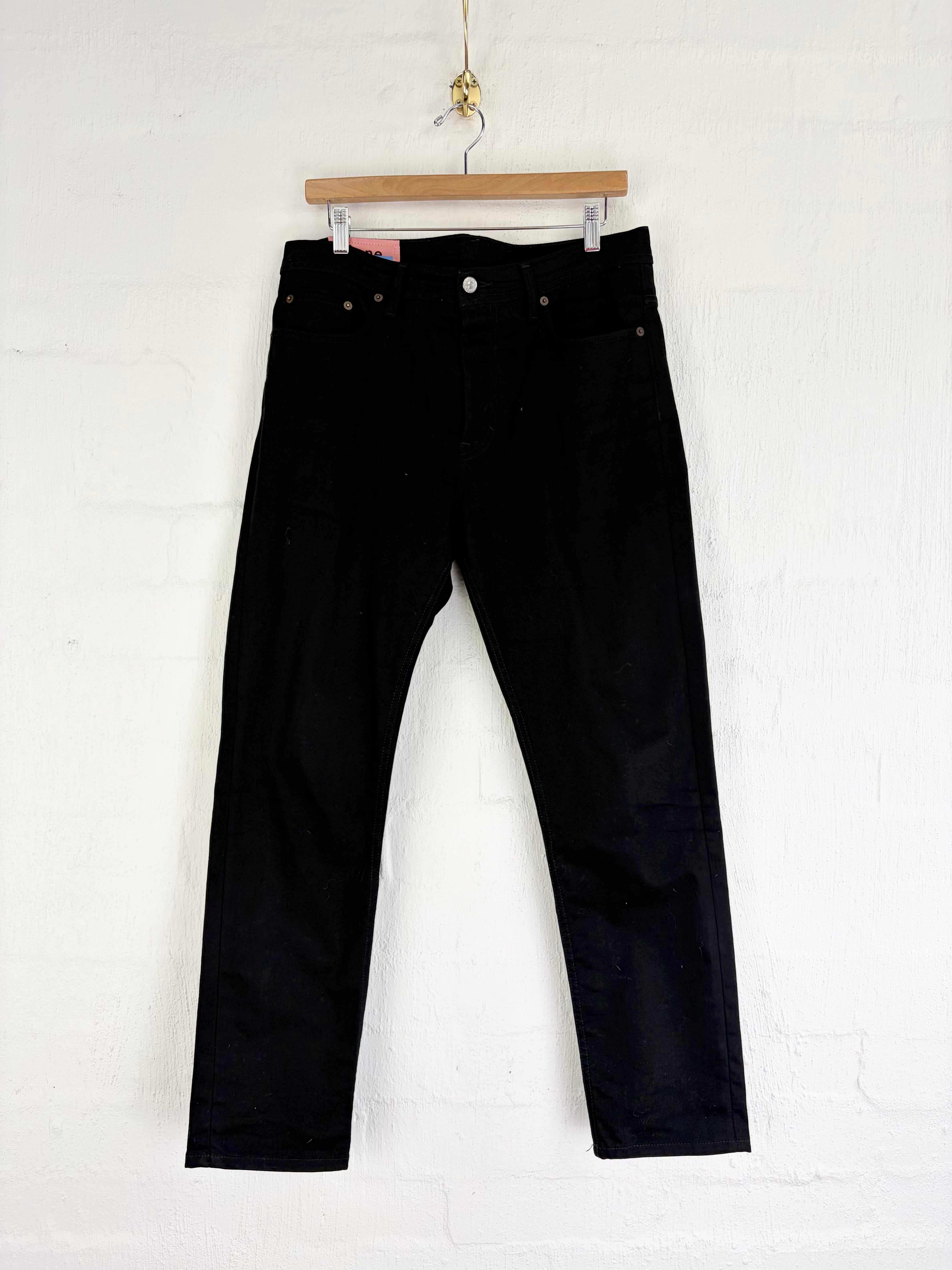 Black denim jeans hanging from a wooden hanger against a white painted brick wall.