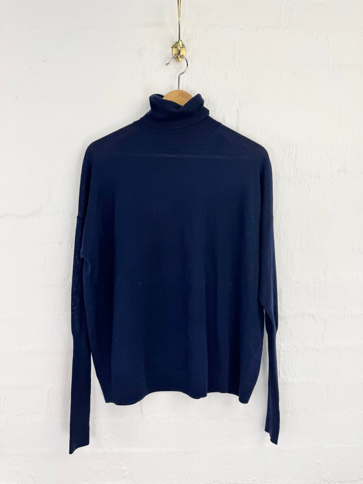 A dark navy blue long-sleeve turtleneck sweater hanging on a wooden hanger against a white painted brick wall.