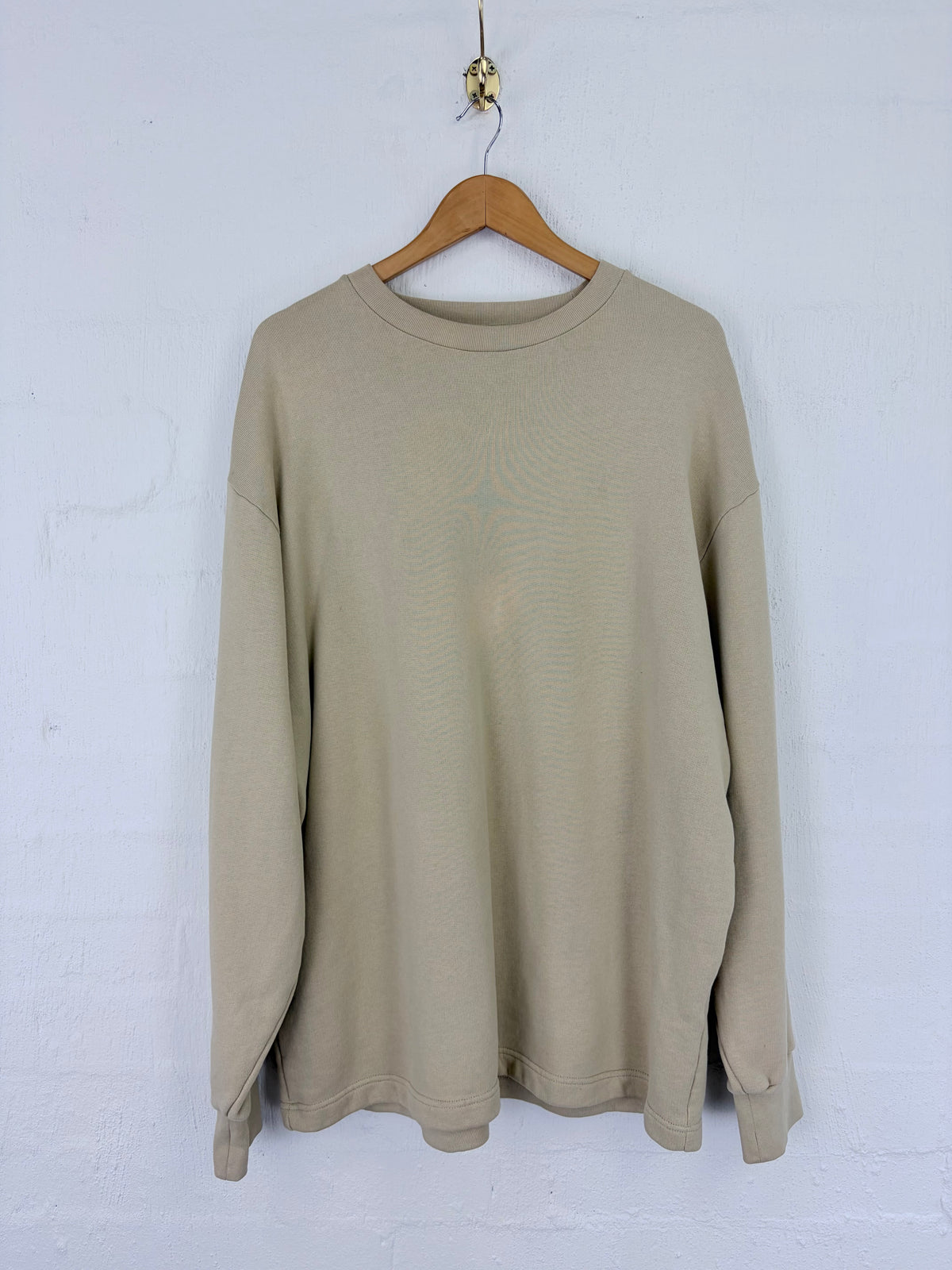 A light beige crewneck sweatshirt hanging on a wooden hanger against a white painted brick wall.