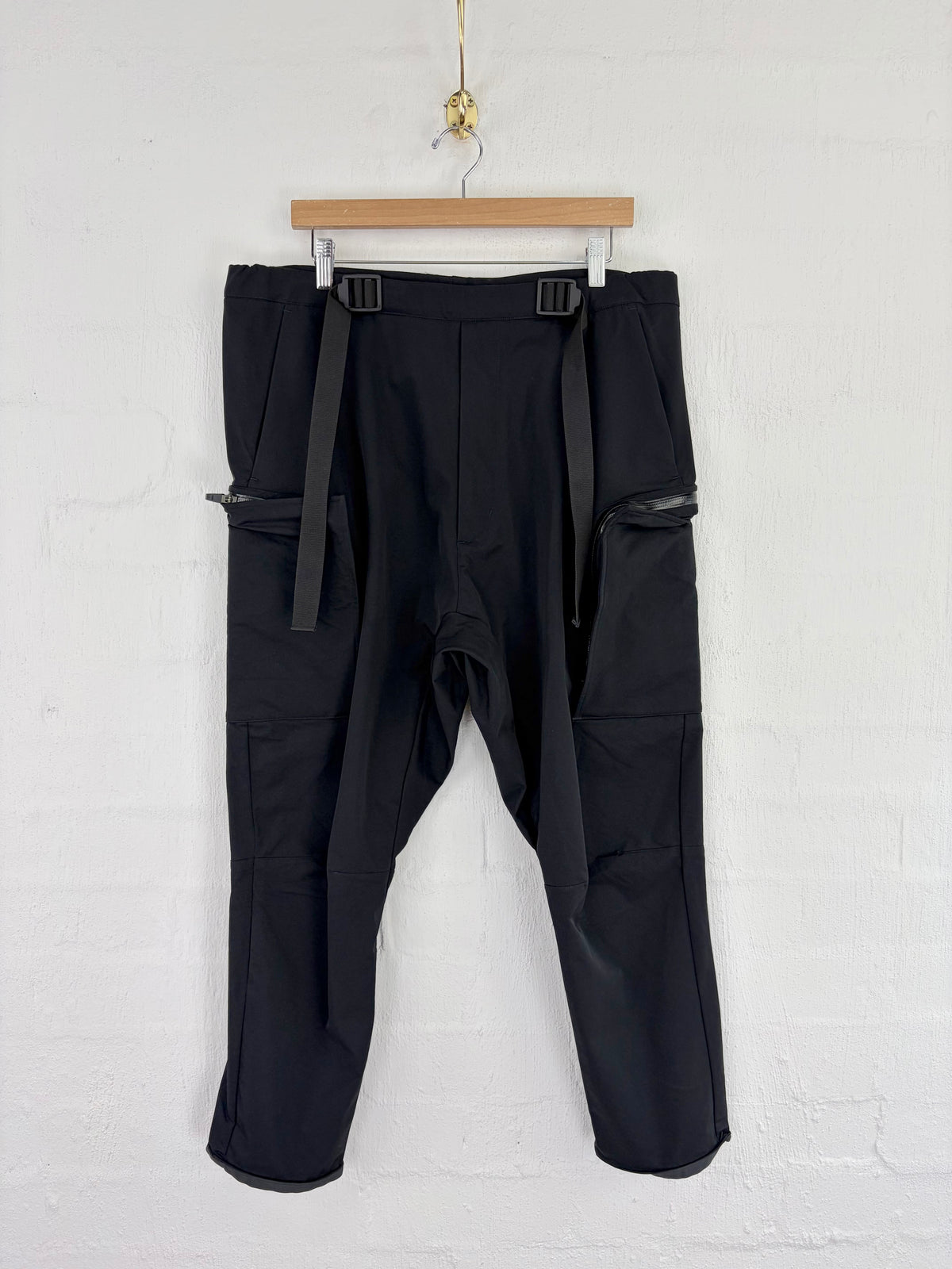Black cargo pants with side straps and zippered pockets, hanging from a wooden hanger against a white brick wall.