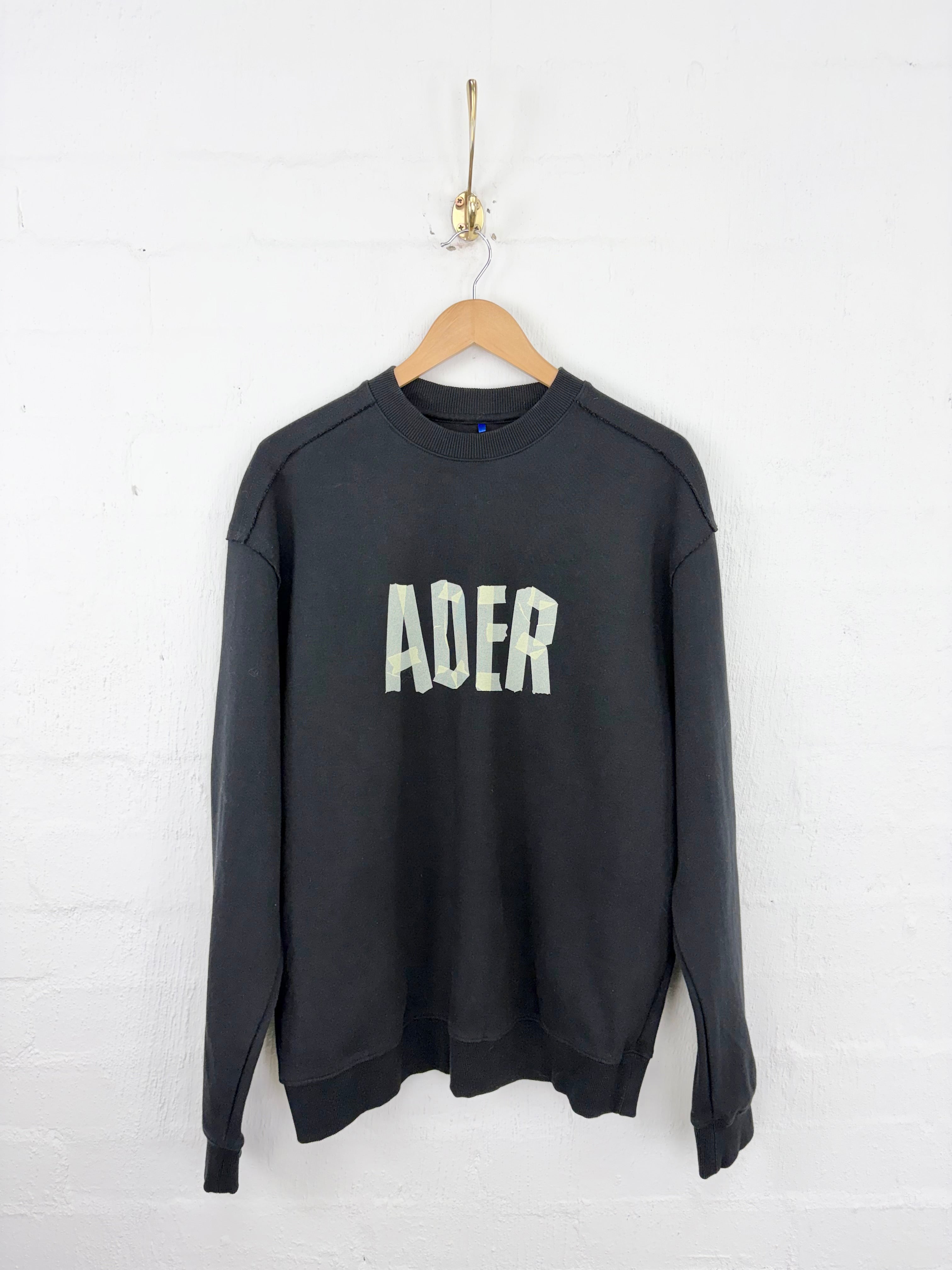 A black long-sleeve crewneck sweatshirt with 'ADER' printed in white across the chest, hanging on a wooden hanger against a white painted brick wall.
