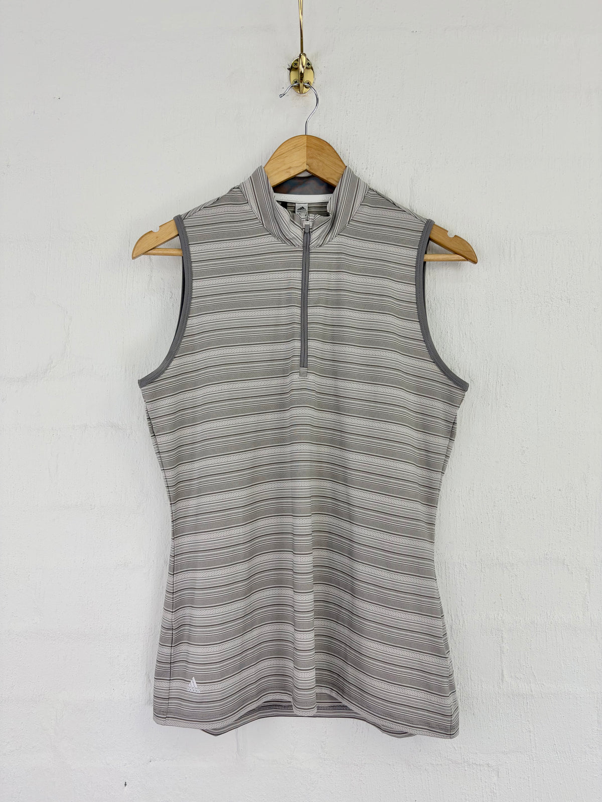 A sleeveless women's athletic top with a quarter-zip collar, featuring a gray and white horizontal stripe pattern, hanging on a wooden hanger against a white brick wall.