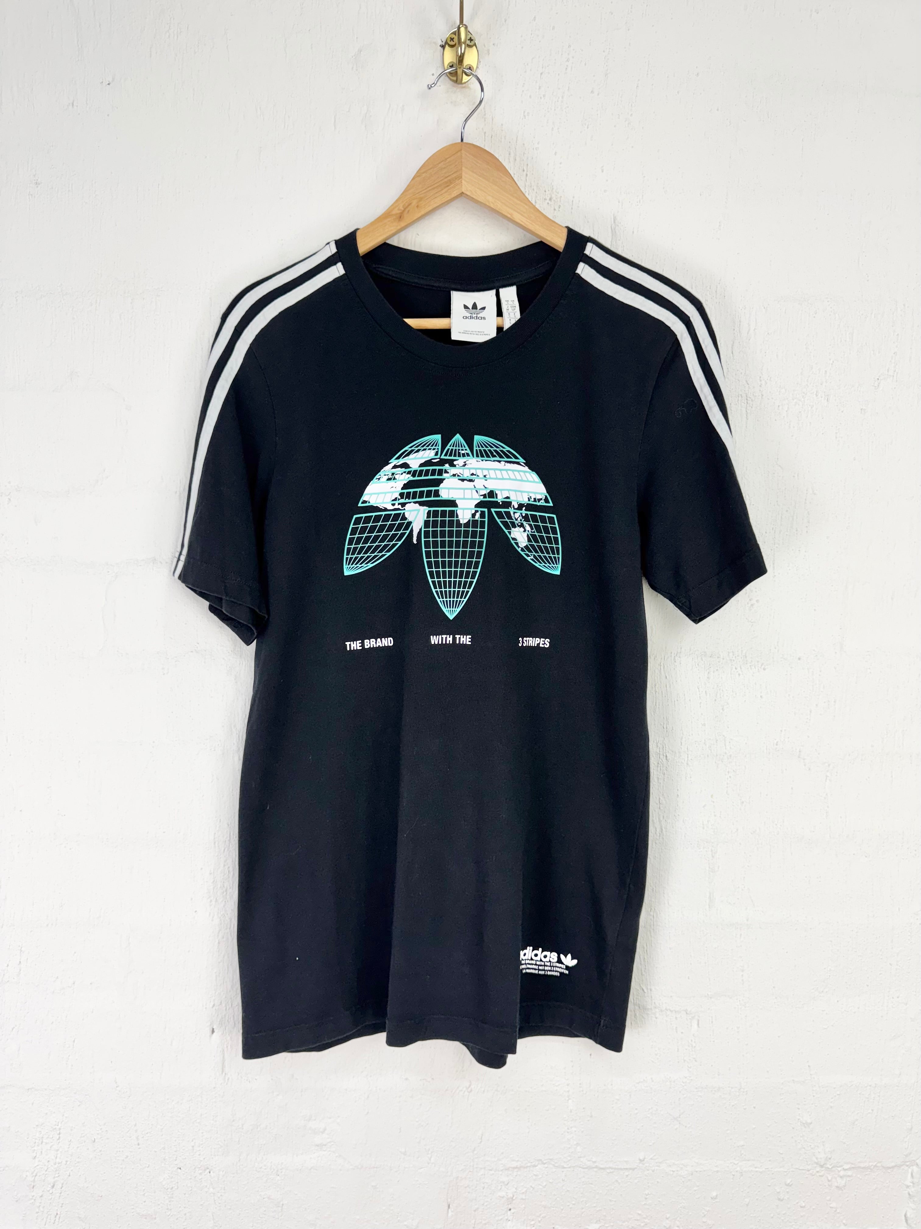 A black Adidas track top with white three-stripes on the sleeves and a green/white globe graphic on the chest, hanging on a wooden hanger against a white brick wall.