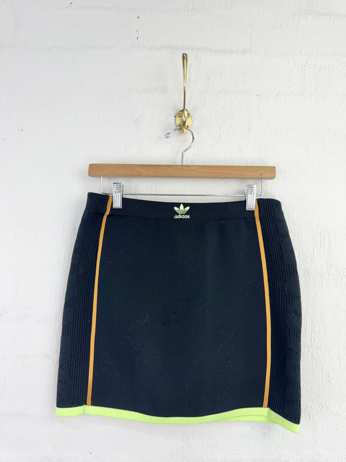 A black Adidas mini skirt with yellow piping and a neon yellow-green hem, hanging on a wooden hanger against a white brick wall.