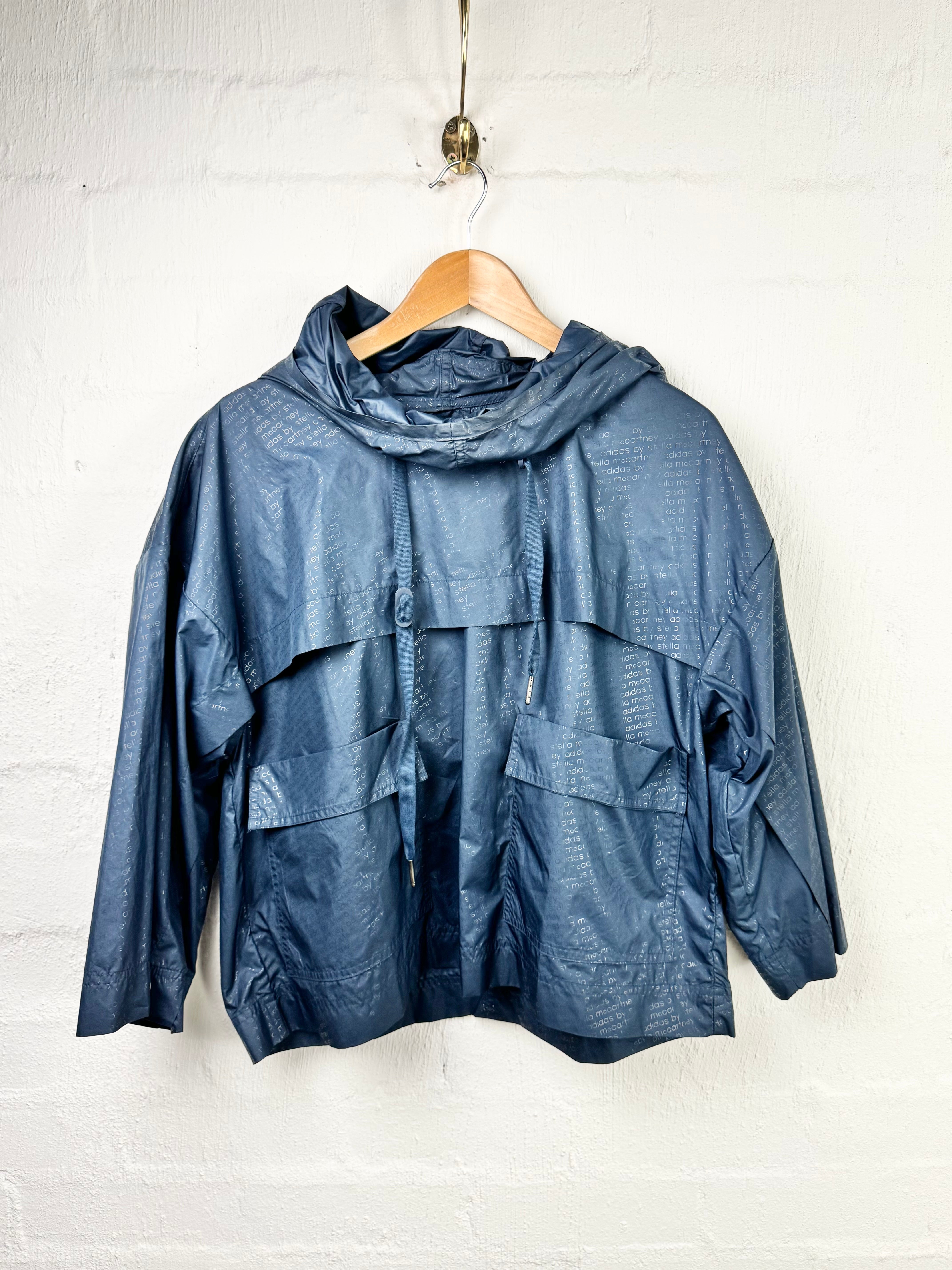 A dark blue hooded rain jacket with a crinkled, textured finish, hanging on a wooden hanger against a white painted brick wall.