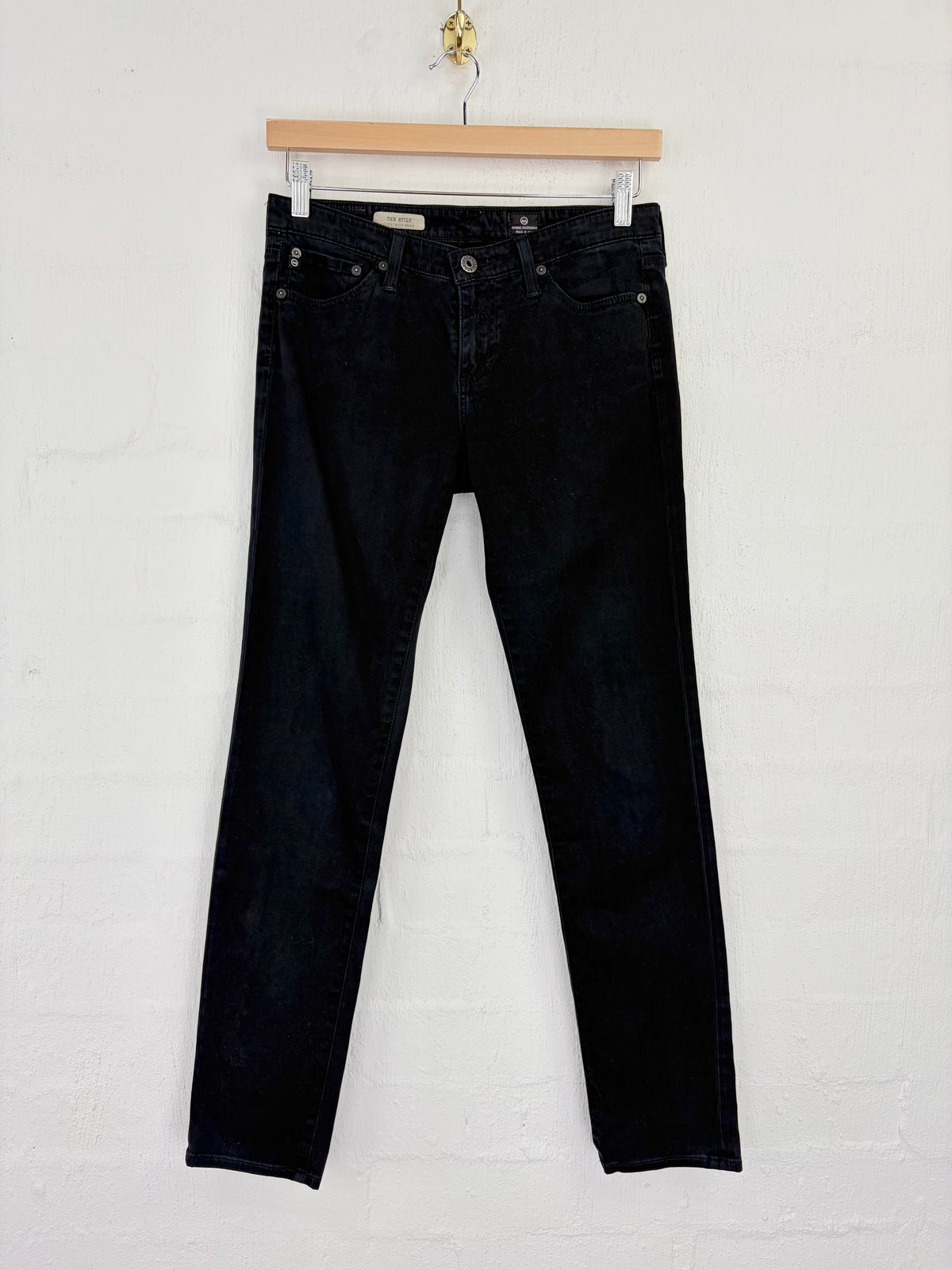 Black denim jeans hanging from a wooden hanger against a white painted brick wall.