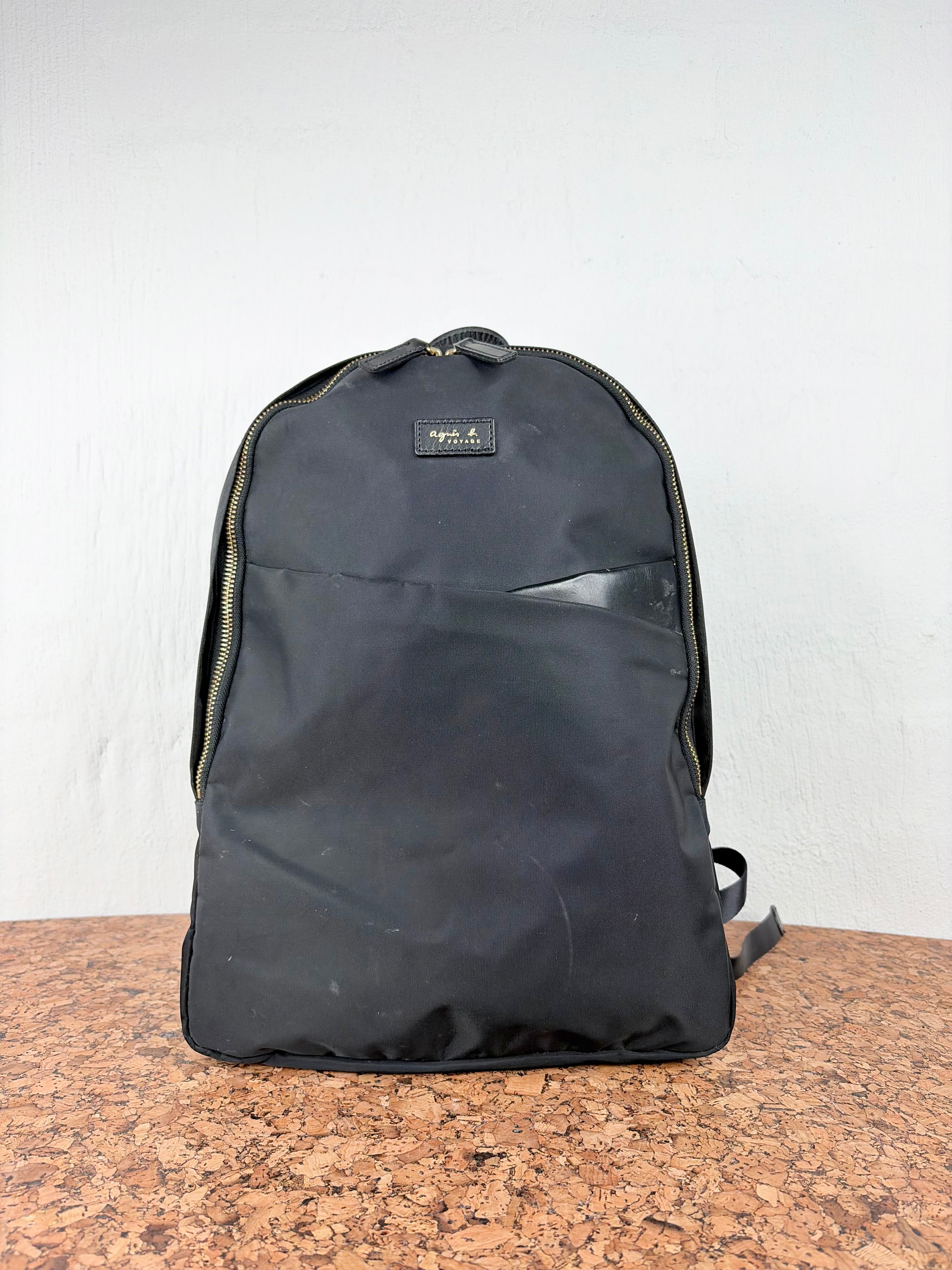 A black nylon backpack with gold-tone zippers and a small metallic brand plaque on the front, standing on a brown cork surface against a white wall.