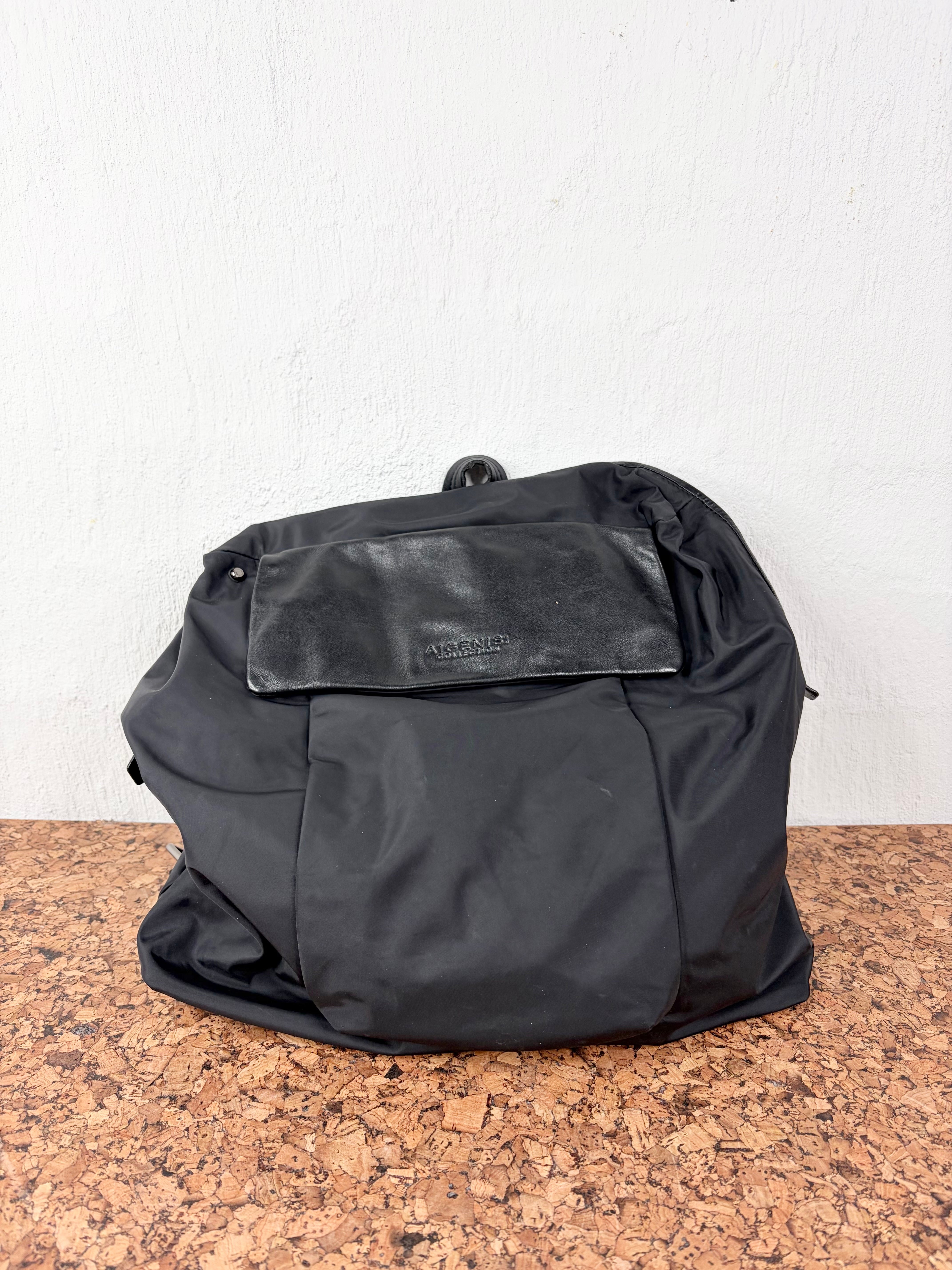 A black nylon backpack with a front flap pocket, resting on a brown cork surface against a white wall.
