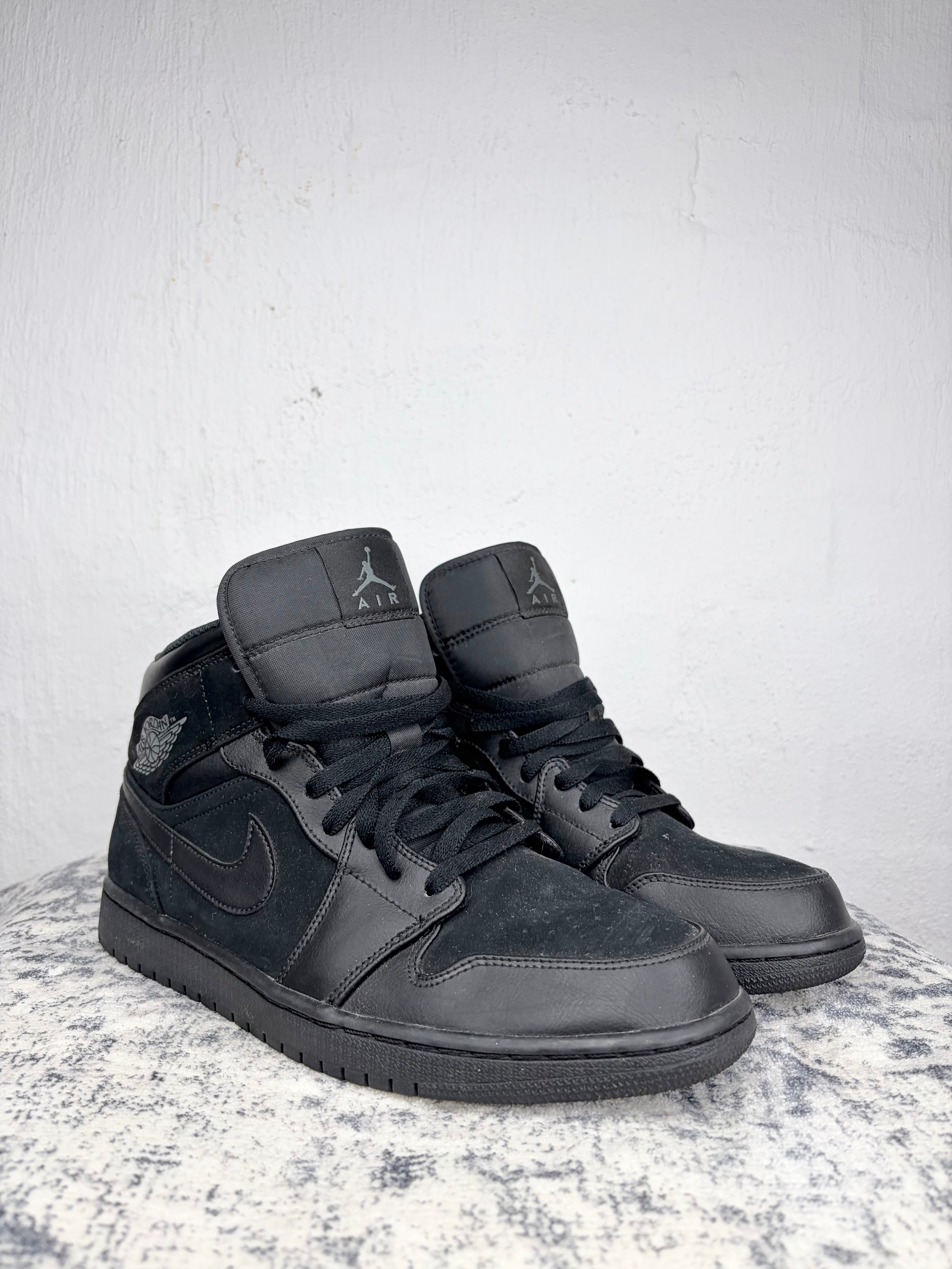 A pair of black Air Jordan 1 Mid sneakers with a mix of smooth leather and suede textures, resting on a gray stone surface against a white textured wall.
