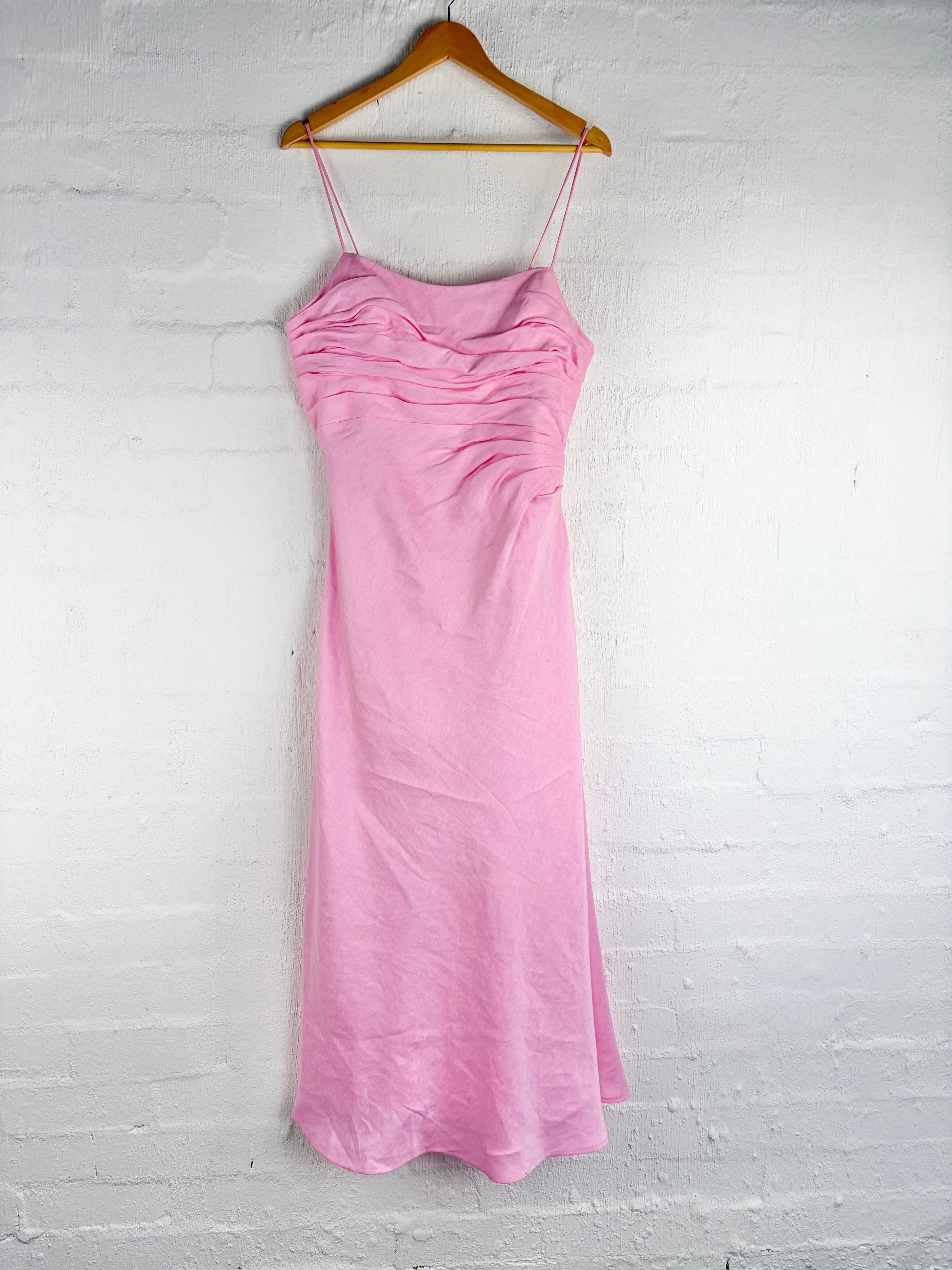 A light pink sleeveless slip dress with thin spaghetti straps and a gathered ruched bodice, hanging from a wooden hanger against a white painted brick wall.