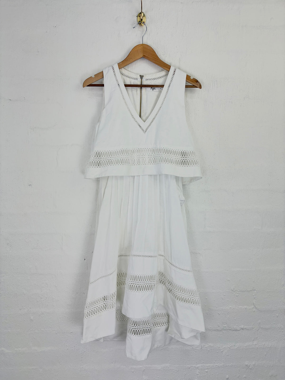 A white sleeveless tiered dress with crochet lace trim details, hanging on a wooden hanger against a white painted brick wall.