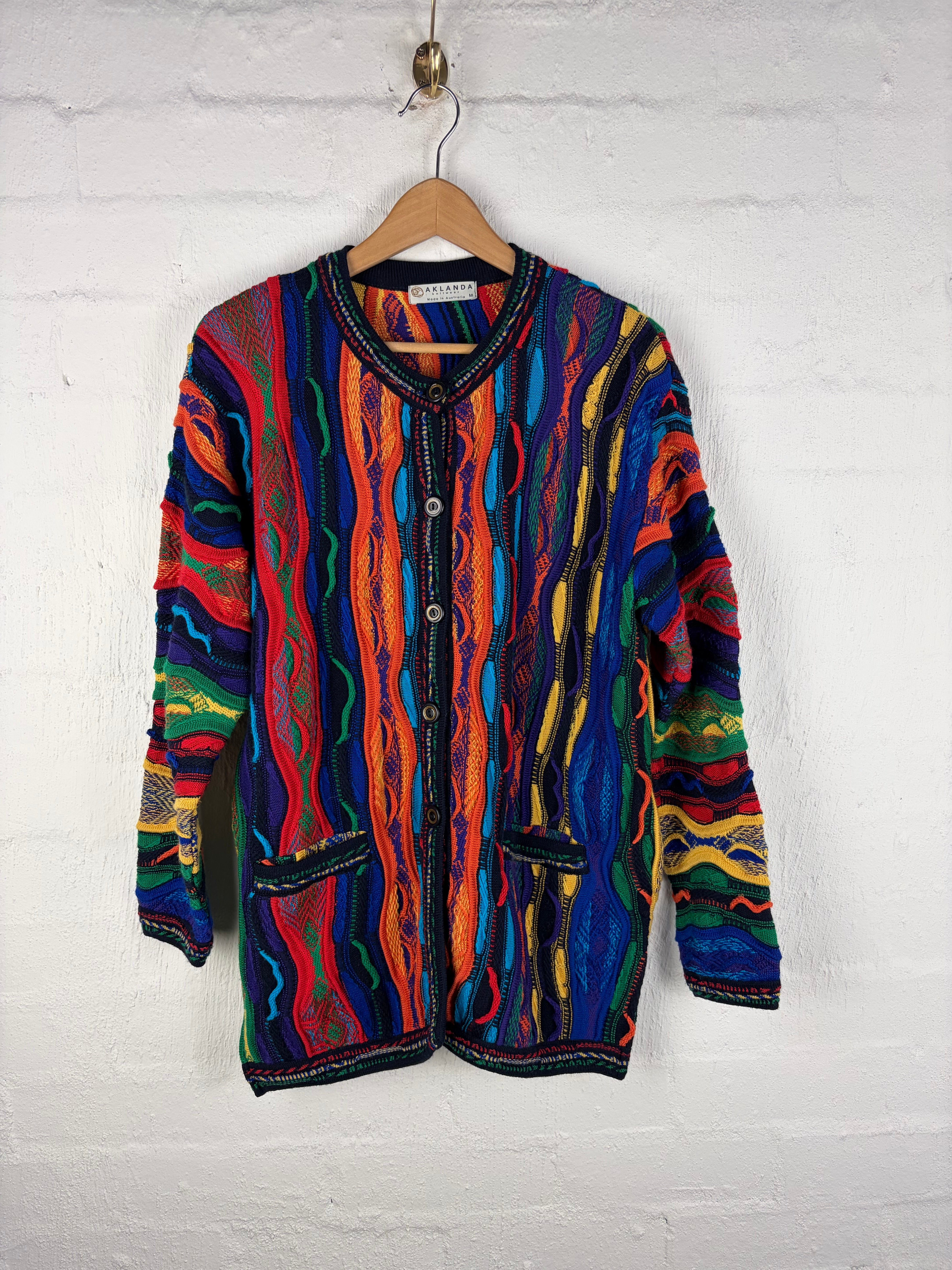 A long-sleeve button-front cardigan with a vibrant, multicolored wavy stripe pattern, hanging on a wooden hanger against a white brick wall.