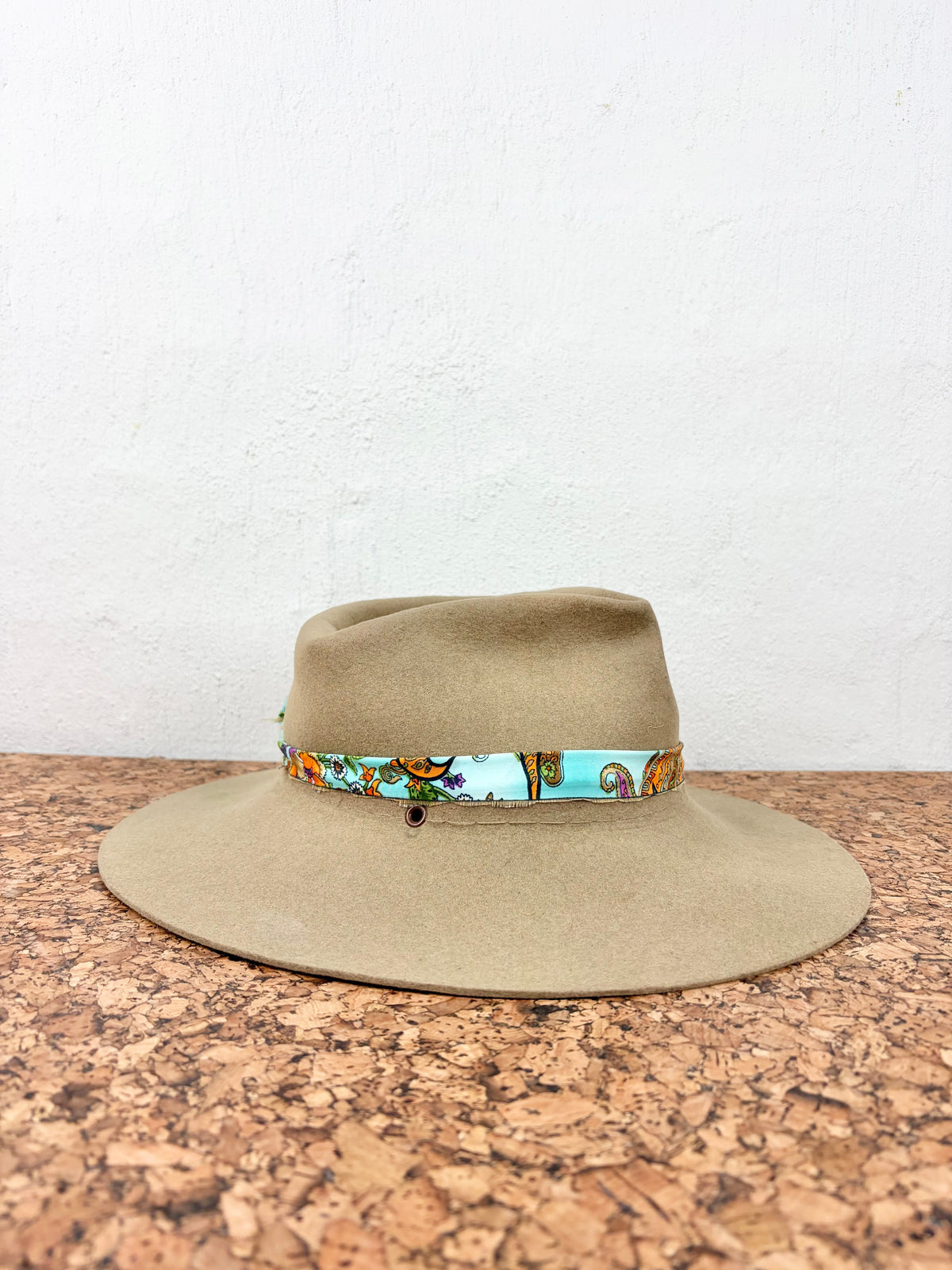 A wide-brimmed tan hat with a colorful patterned band around the crown, resting on a cork surface against a white wall.