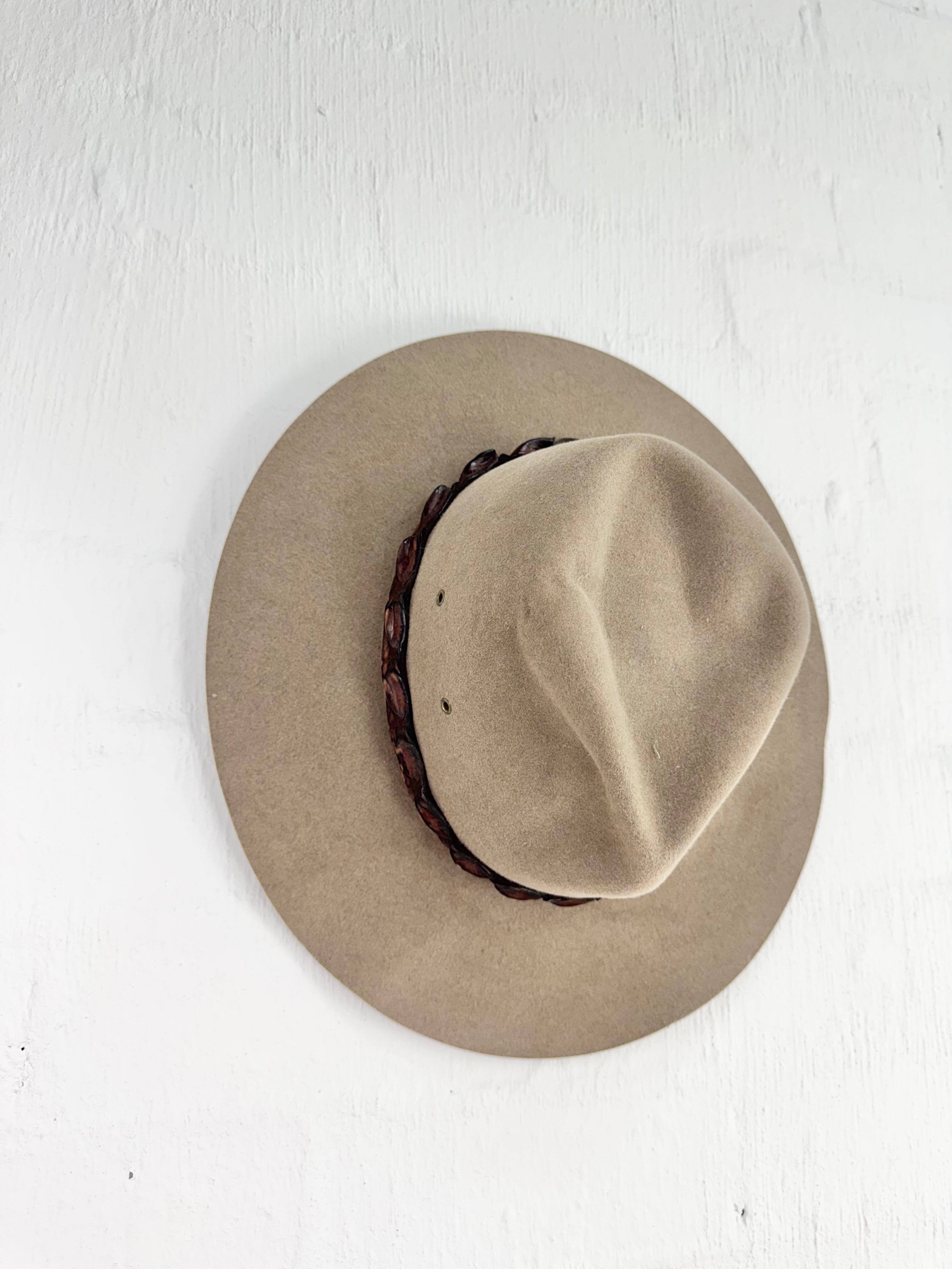 A light brown felt wide-brim hat with a dark brown braided band, photographed from a top-down perspective on a white textured background.