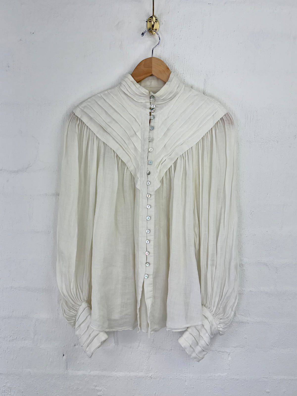 An off-white long-sleeve blouse with a high pleated mock neck and gathered cuffs, hanging on a wooden hanger against a white brick wall.