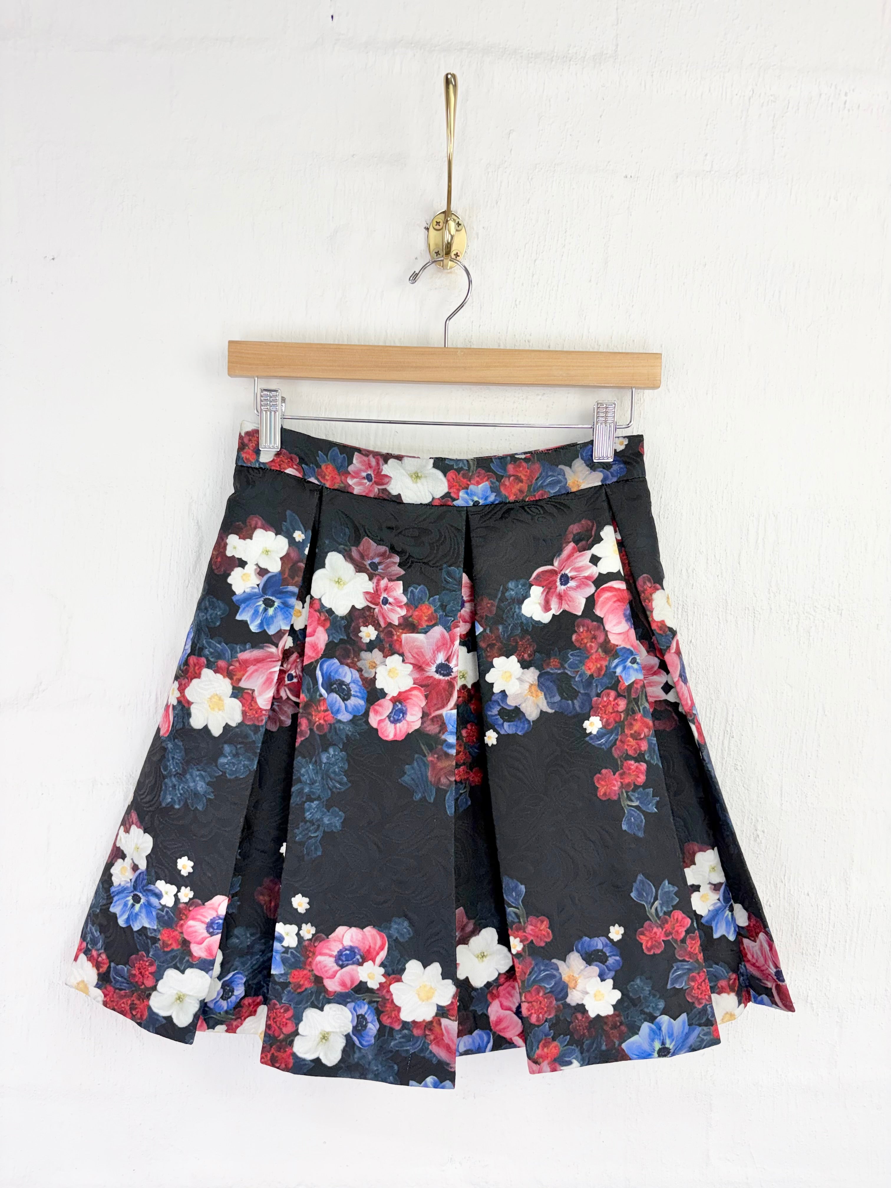 A black pleated skirt with a vibrant multicolor floral print, hanging from a wooden hanger against a white textured wall.