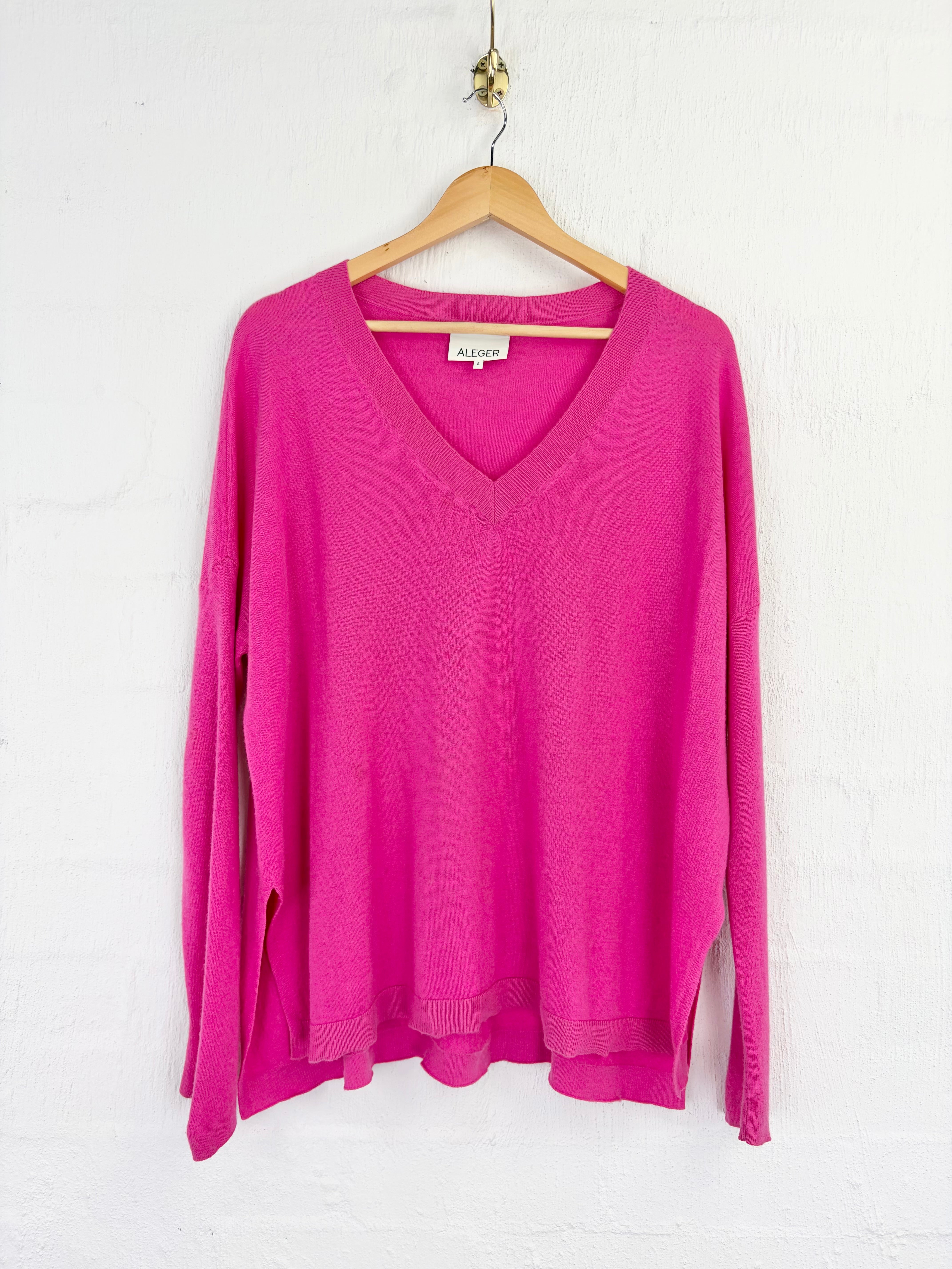 A bright pink long-sleeve V-neck knit sweater hanging on a wooden hanger against a white textured wall.