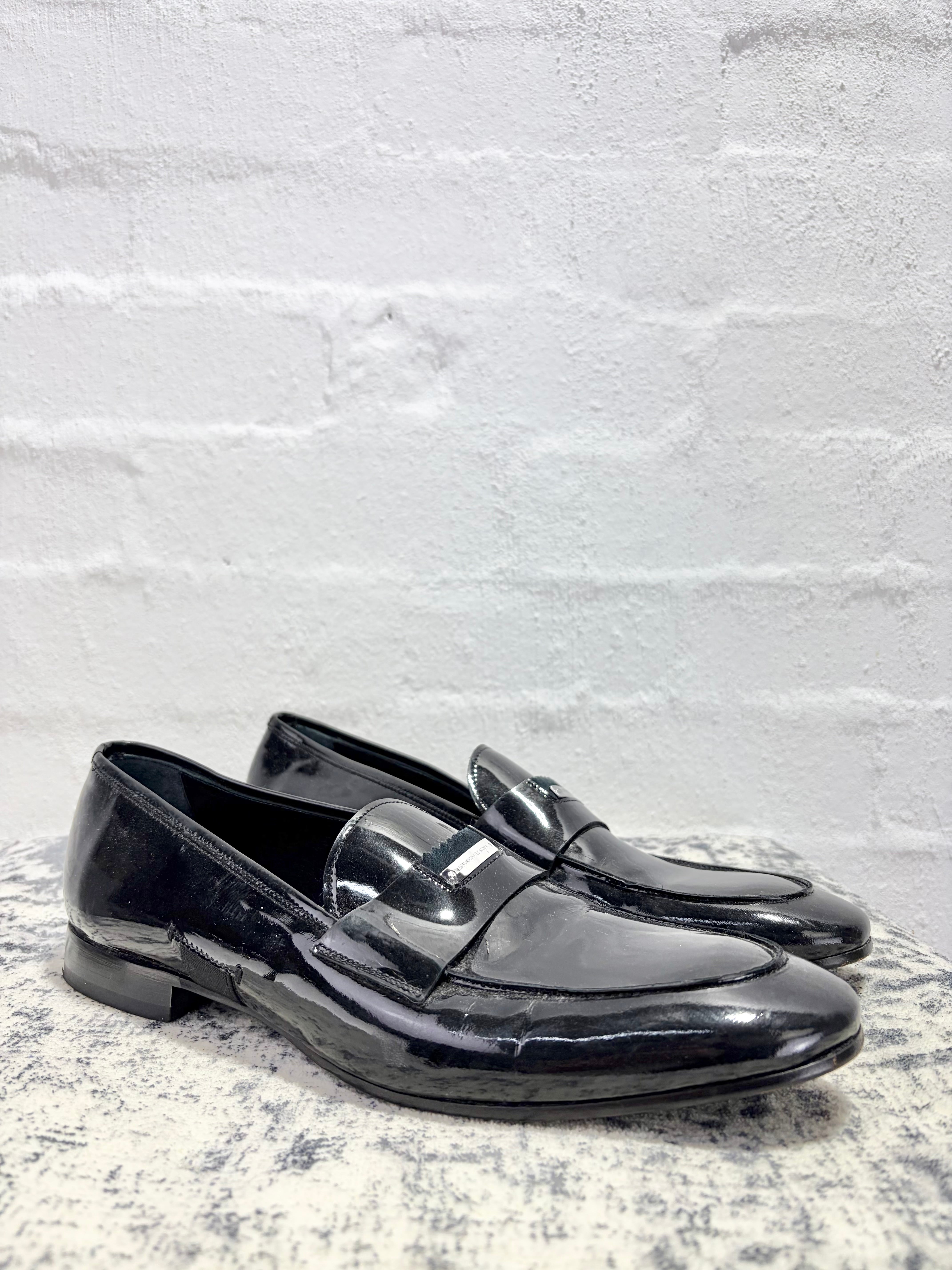 A pair of glossy black patent leather penny loafers resting on a textured gray stone surface against a white brick wall background.