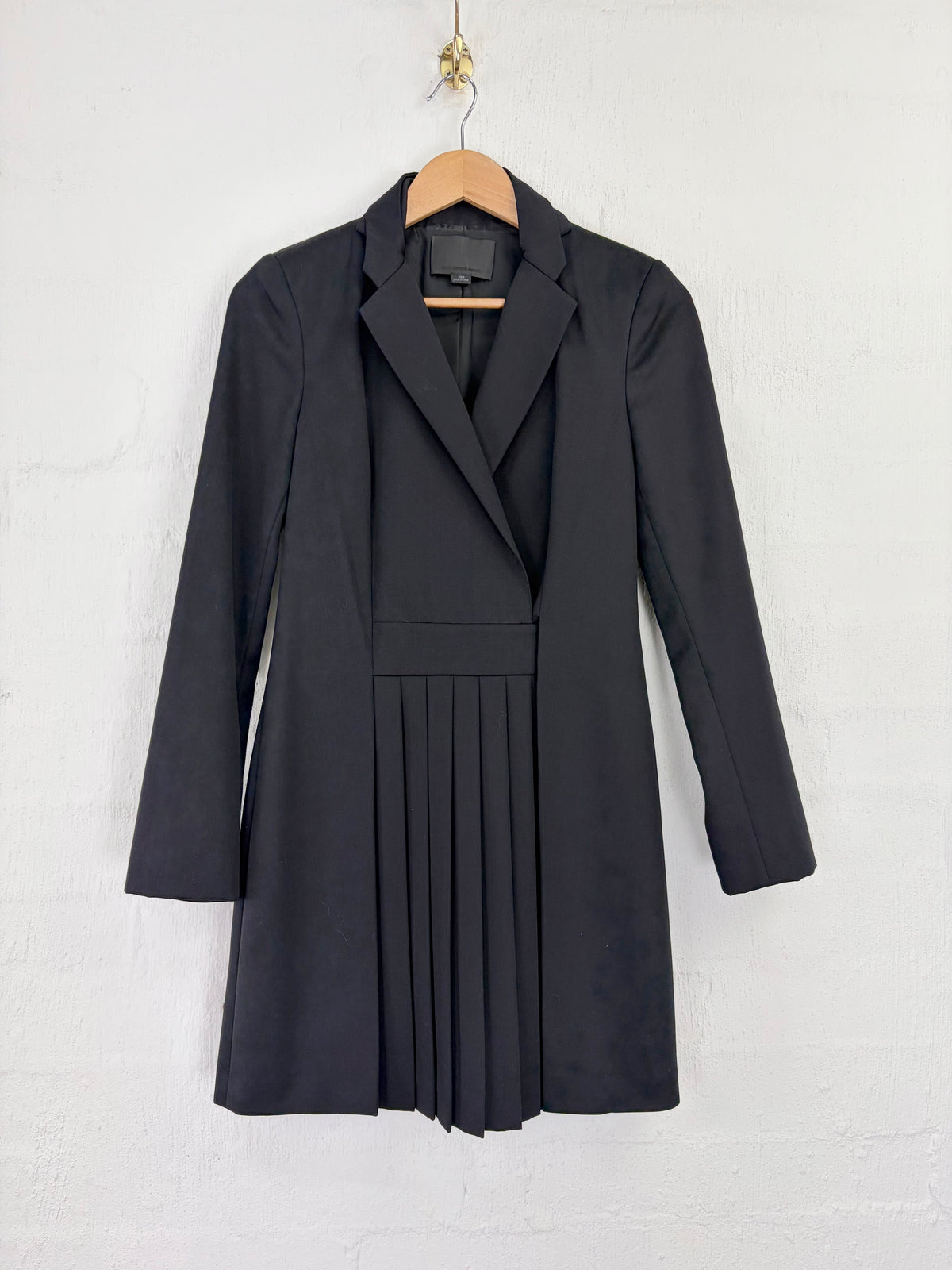A black long-sleeve blazer dress with a pleated skirt detail, hanging on a wooden hanger against a white textured wall.