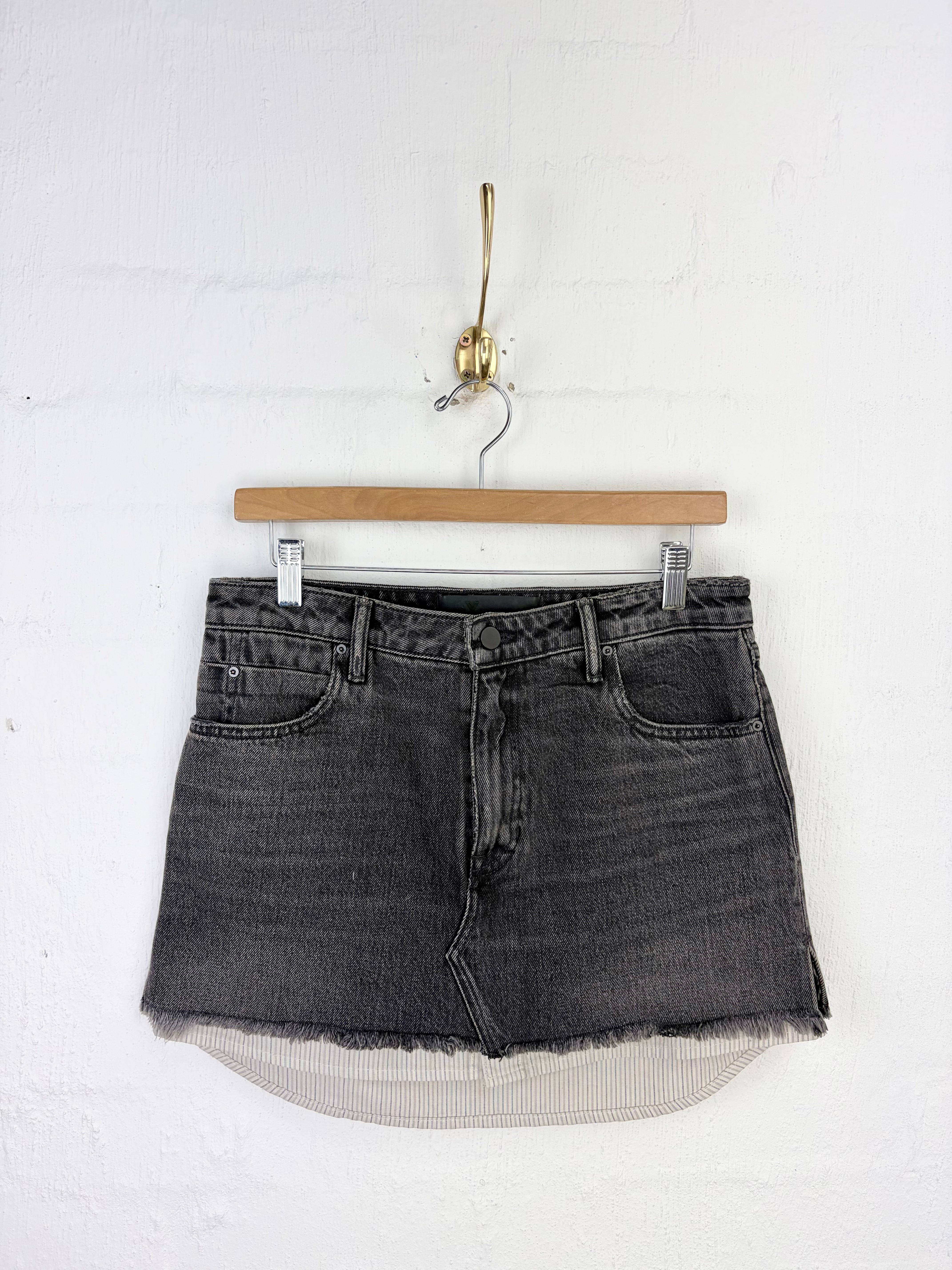 A dark gray washed denim mini skirt with a raw, frayed hem, hanging from a wooden hanger on a white brick wall.