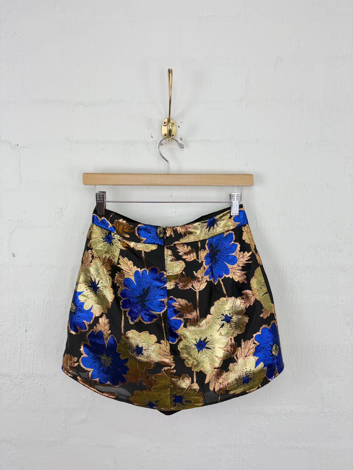 A high-waisted mini skirt with a metallic gold and blue floral pattern on a black base, hanging from a wooden hanger against a white brick wall.